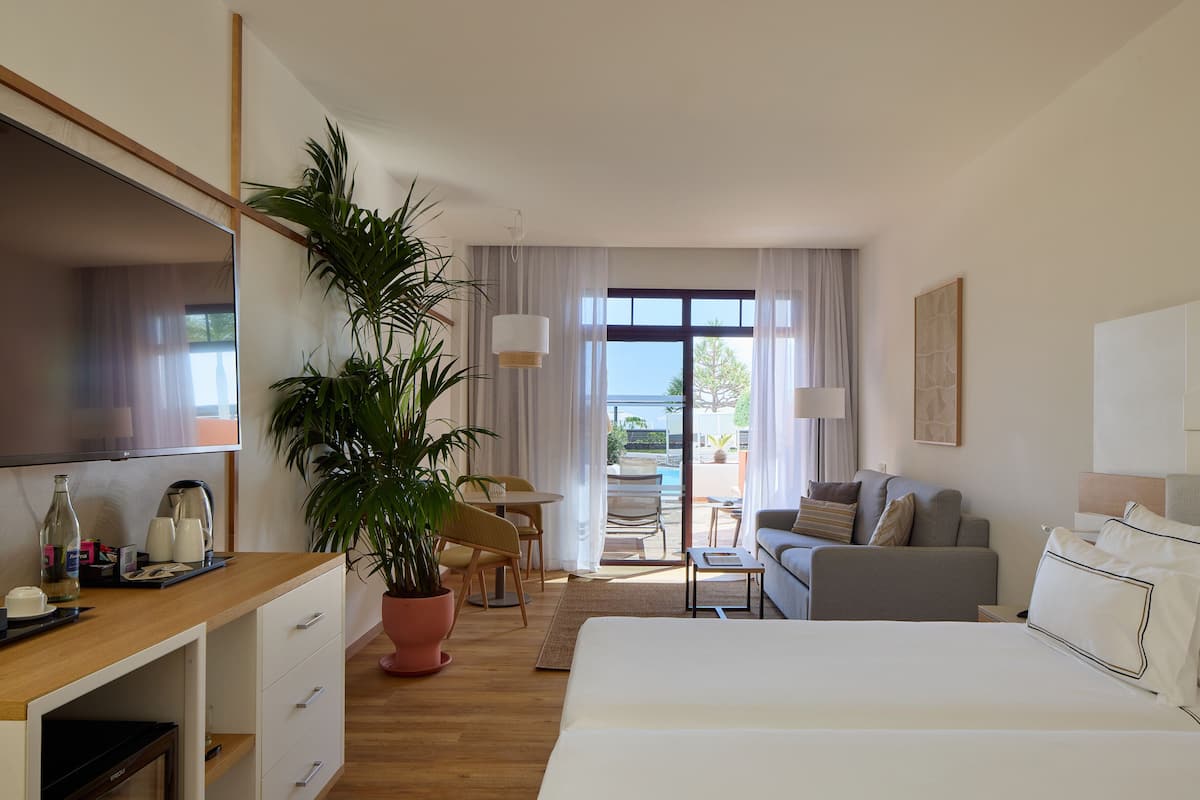 A hotel room with a bed, a desk with a TV, a dining area with a table and chairs, and a large window with curtains.