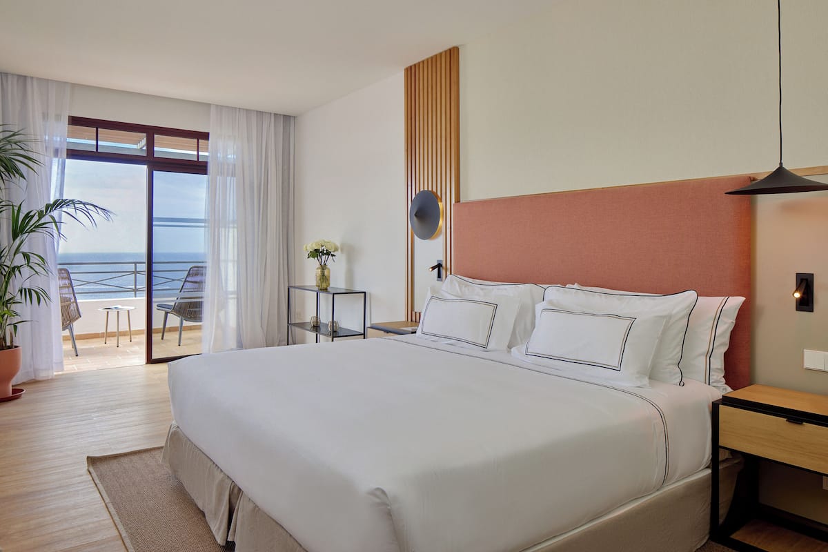 A hotel room with a large bed, a small table, a chair, a potted plant, and a view of the sea through a sliding door.