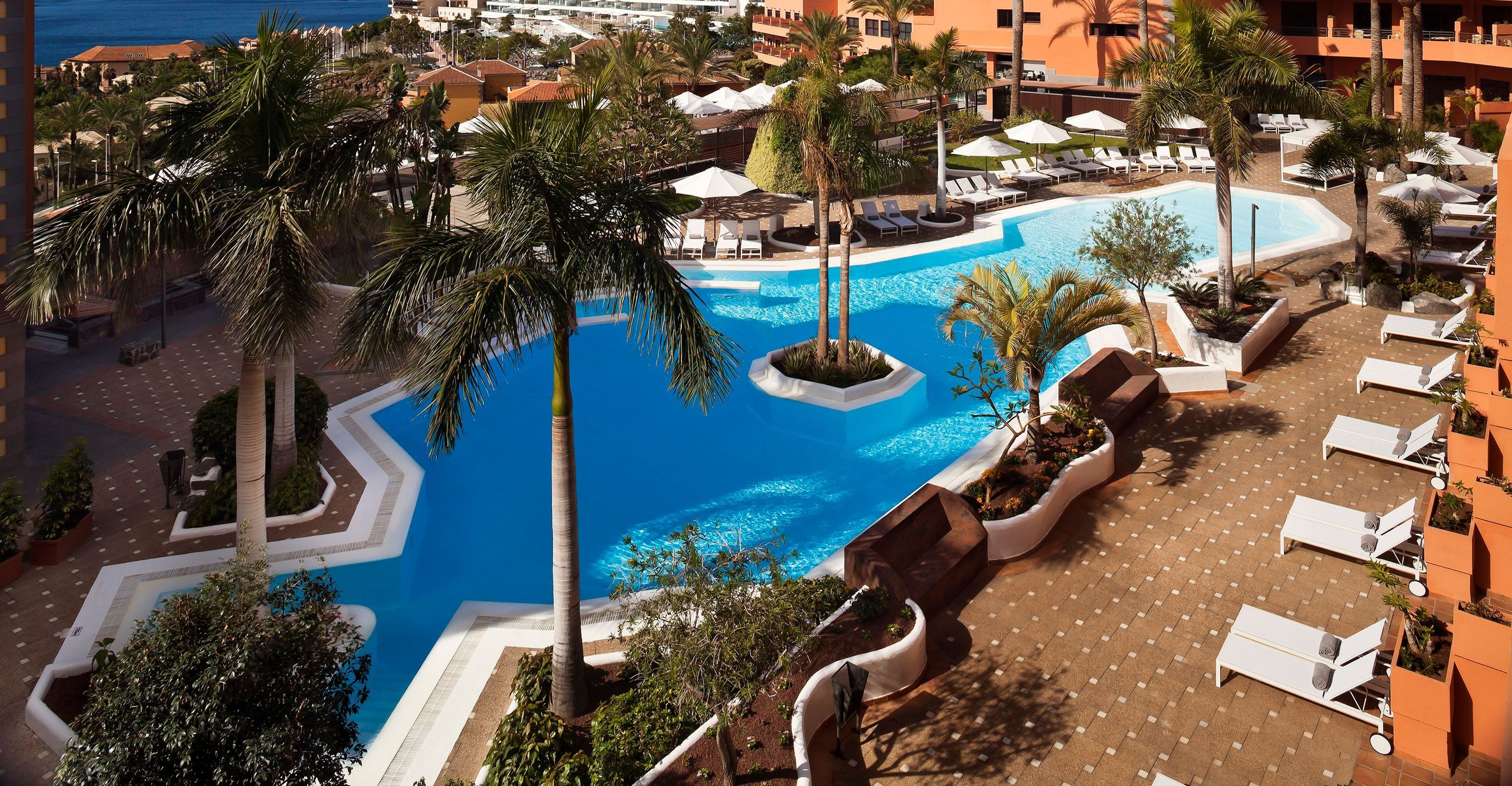 Un área de piscina en un resort con sillas de descanso, palmeras y una piscina azul cristalina.