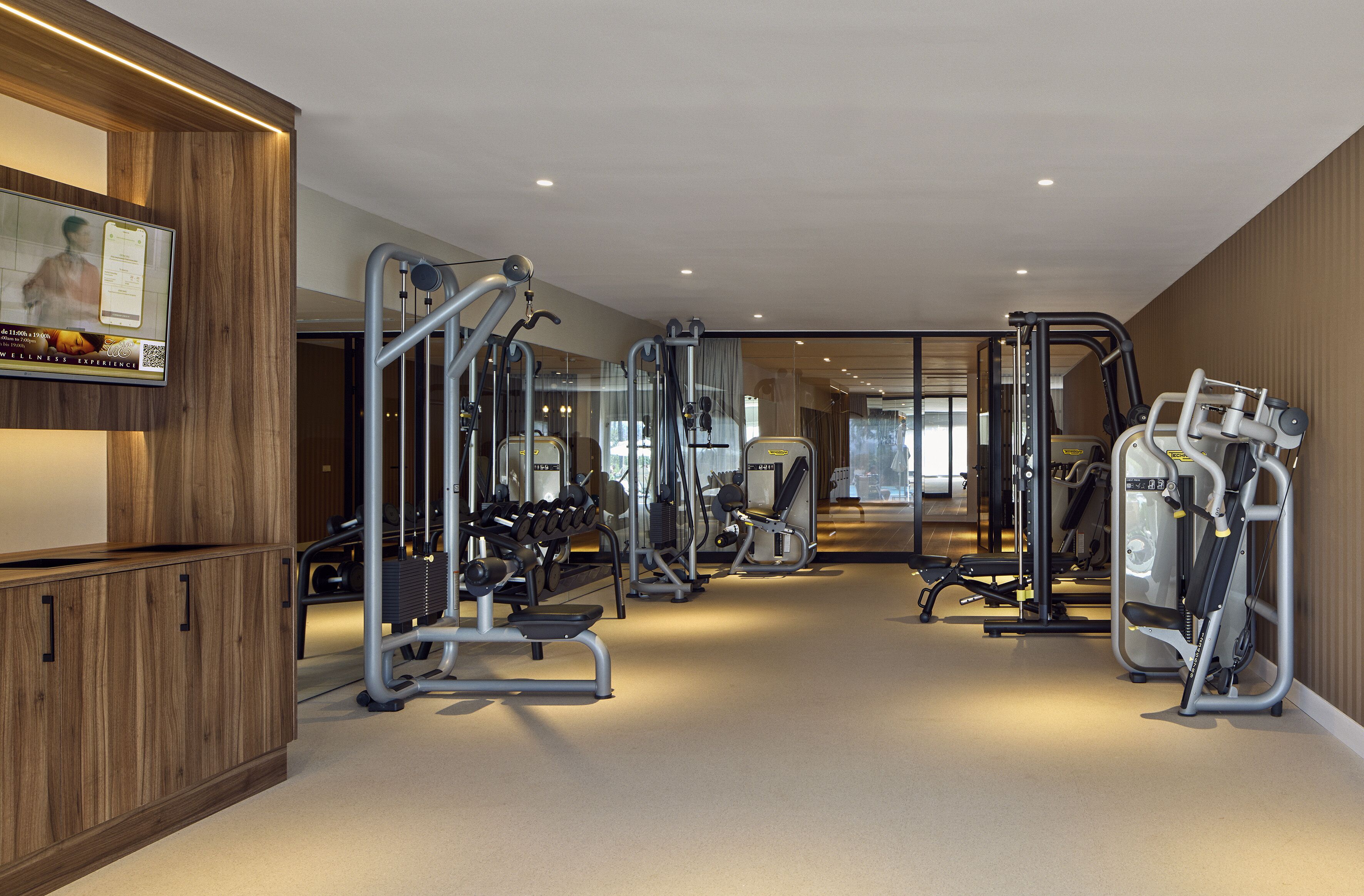 Un gimnasio moderno con diversas máquinas de ejercicio, un mostrador de recepción de madera y un espejo grande que refleja el espacio.