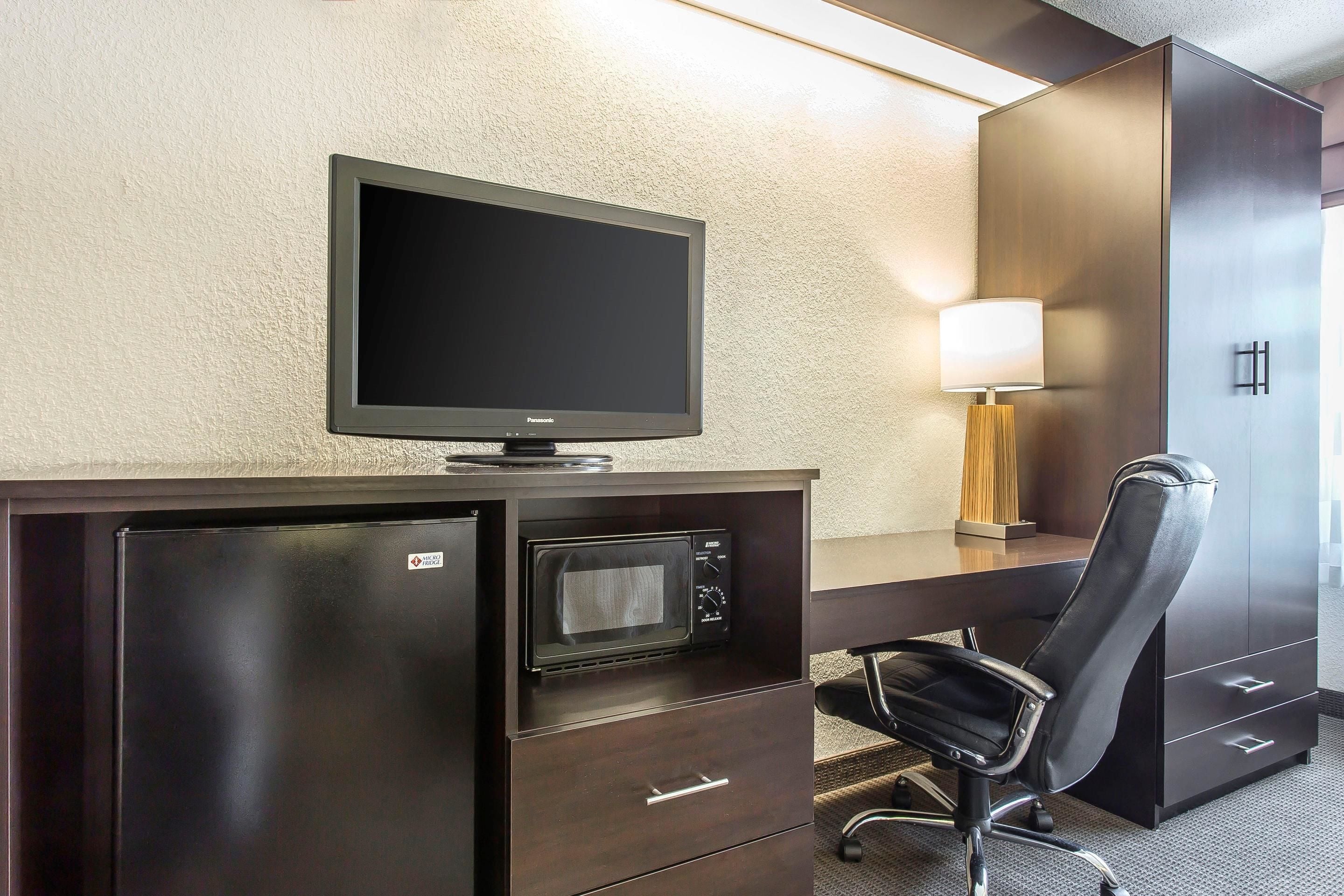 A hotel room with a dark wooden desk, a flat-screen TV, a microwave, a lamp, a chair, and a minibar.