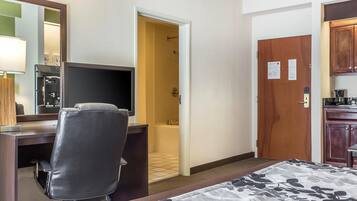 A hotel room with a bed, a desk with a chair, a television, a bathroom with a shower, and a wooden cabinet.