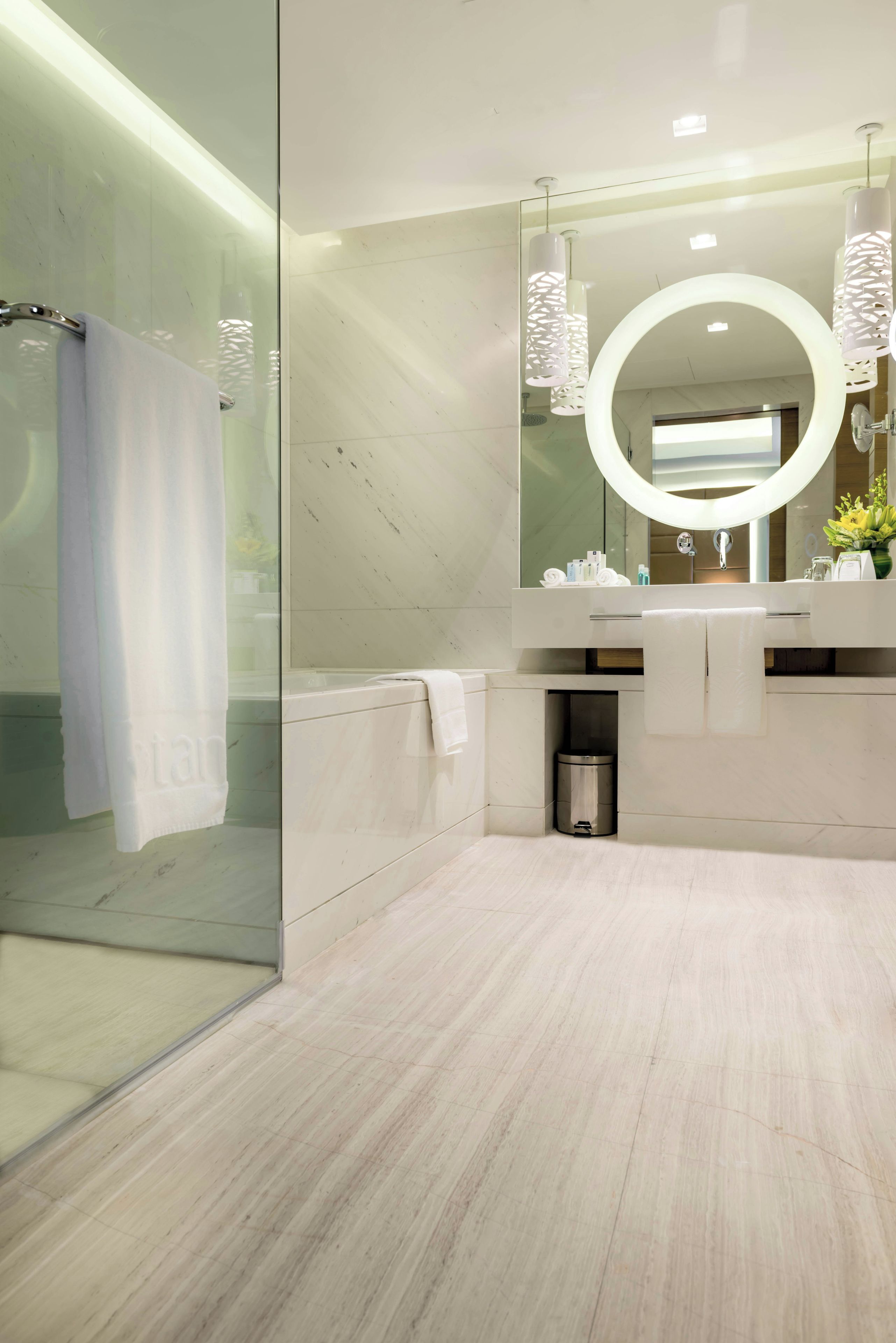 A modern bathroom with a glass shower enclosure, a marble-tiled floor, a large mirror with a circular frame, and a white towel hanging on the glass door.