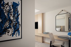 A modern bedroom with a large abstract painting, a desk with a computer, a chair, and a bedside table with a vase of flowers.