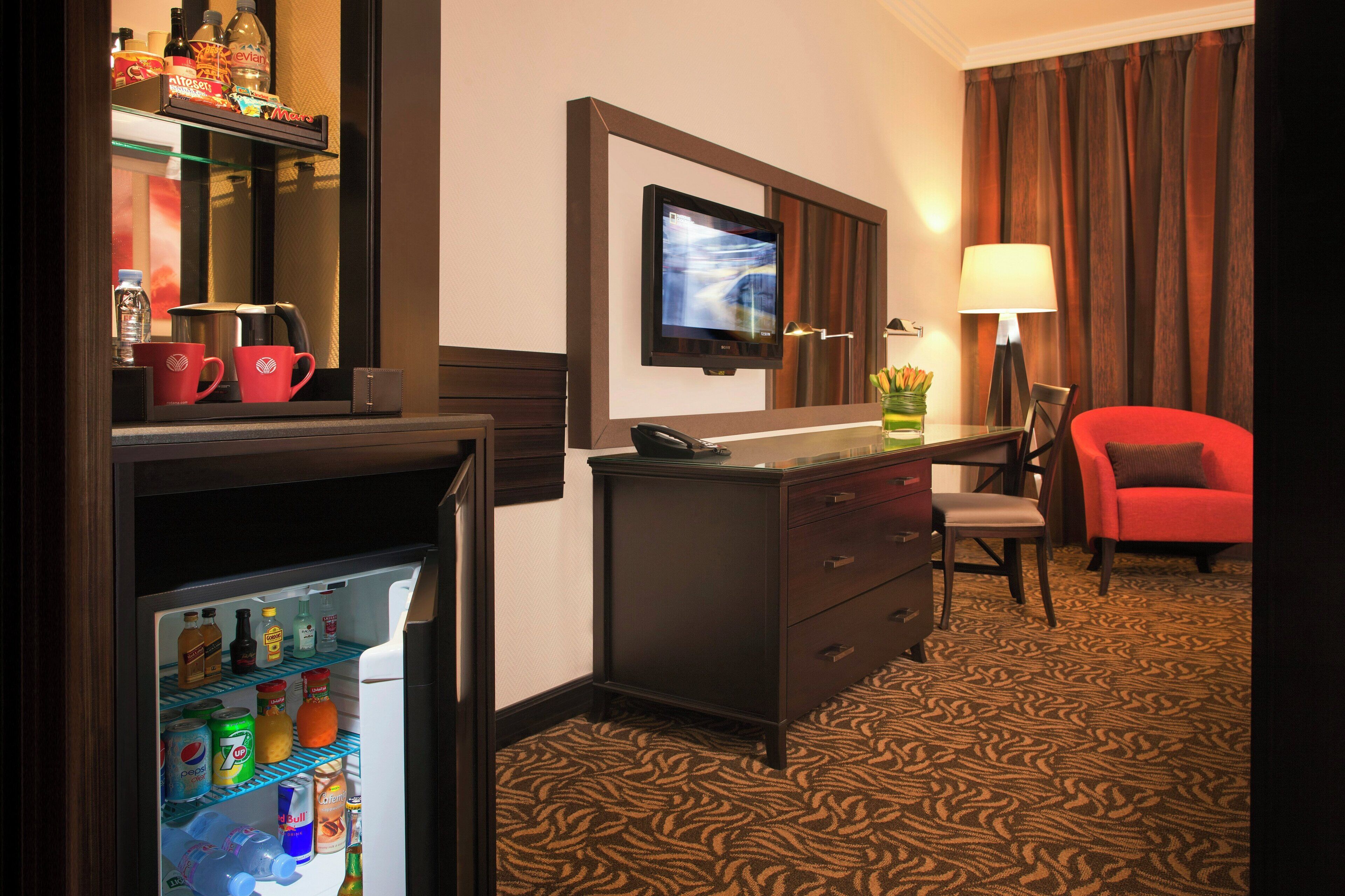 A hotel room with a minibar, a television, a desk with a chair, and a red sofa.