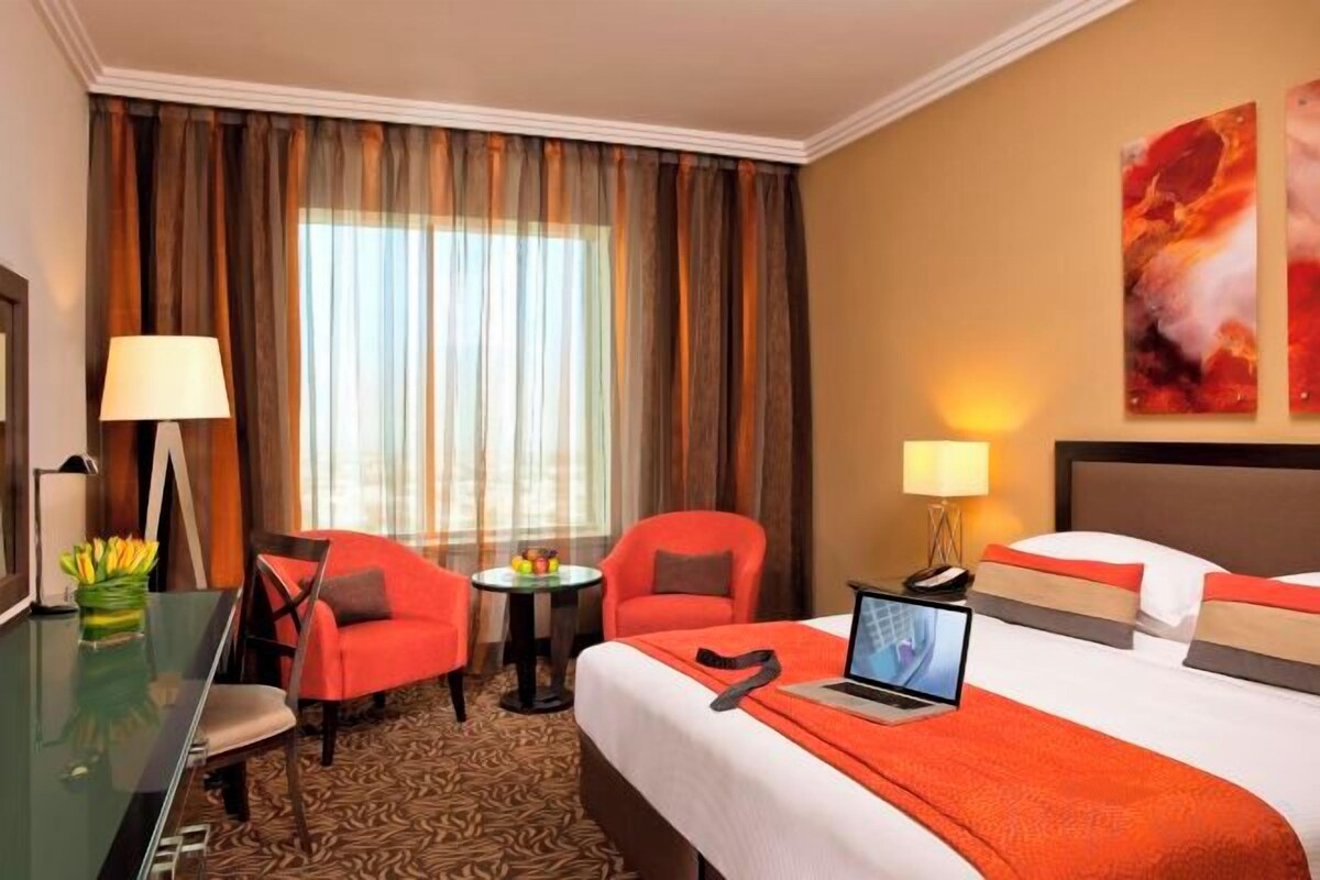 A hotel room with a bed, two chairs, a desk, a laptop, a lamp, and a view of the outdoors.