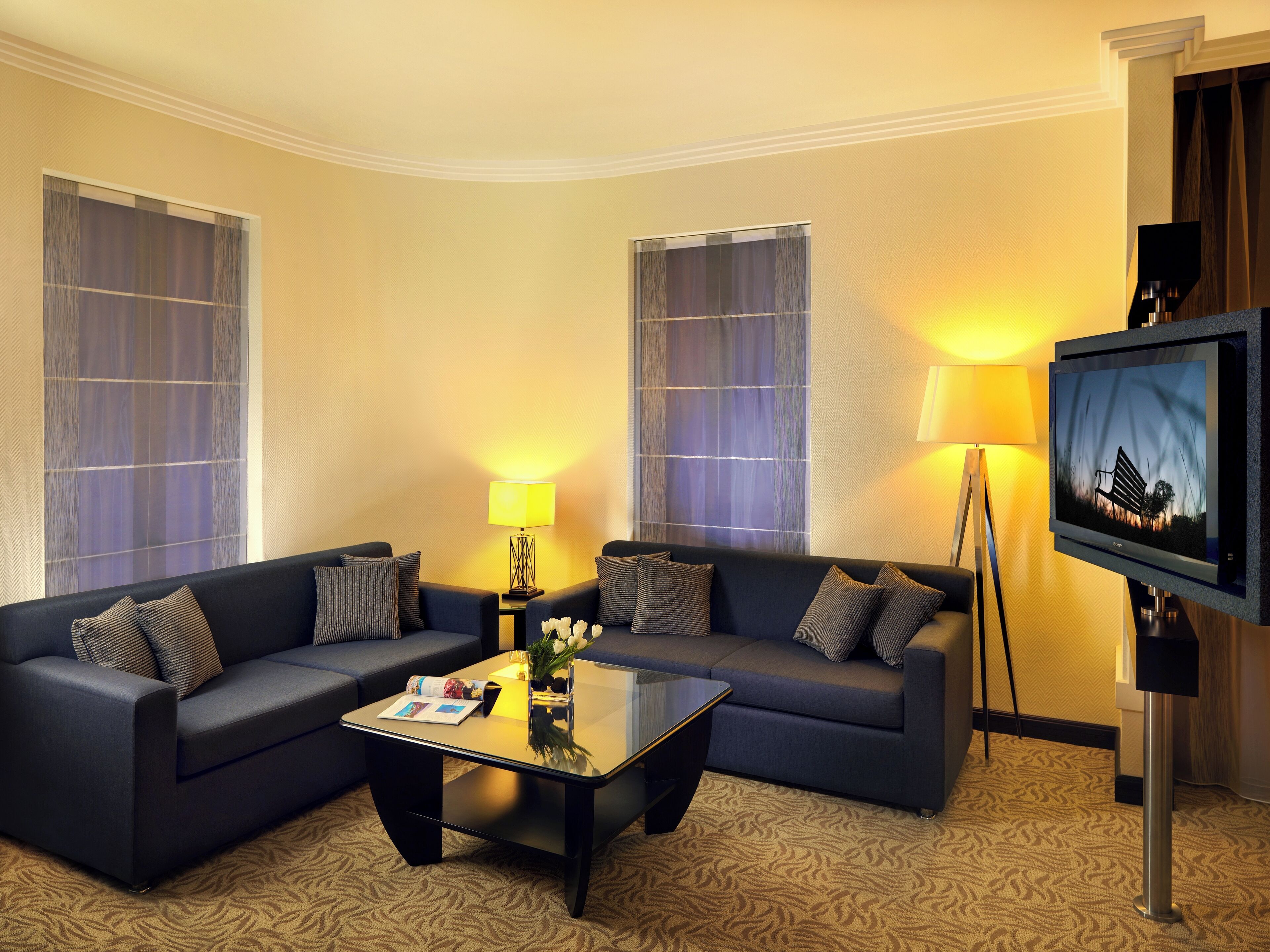 A hotel room with two dark blue sofas, a glass coffee table, and a flat-screen TV mounted on the wall.