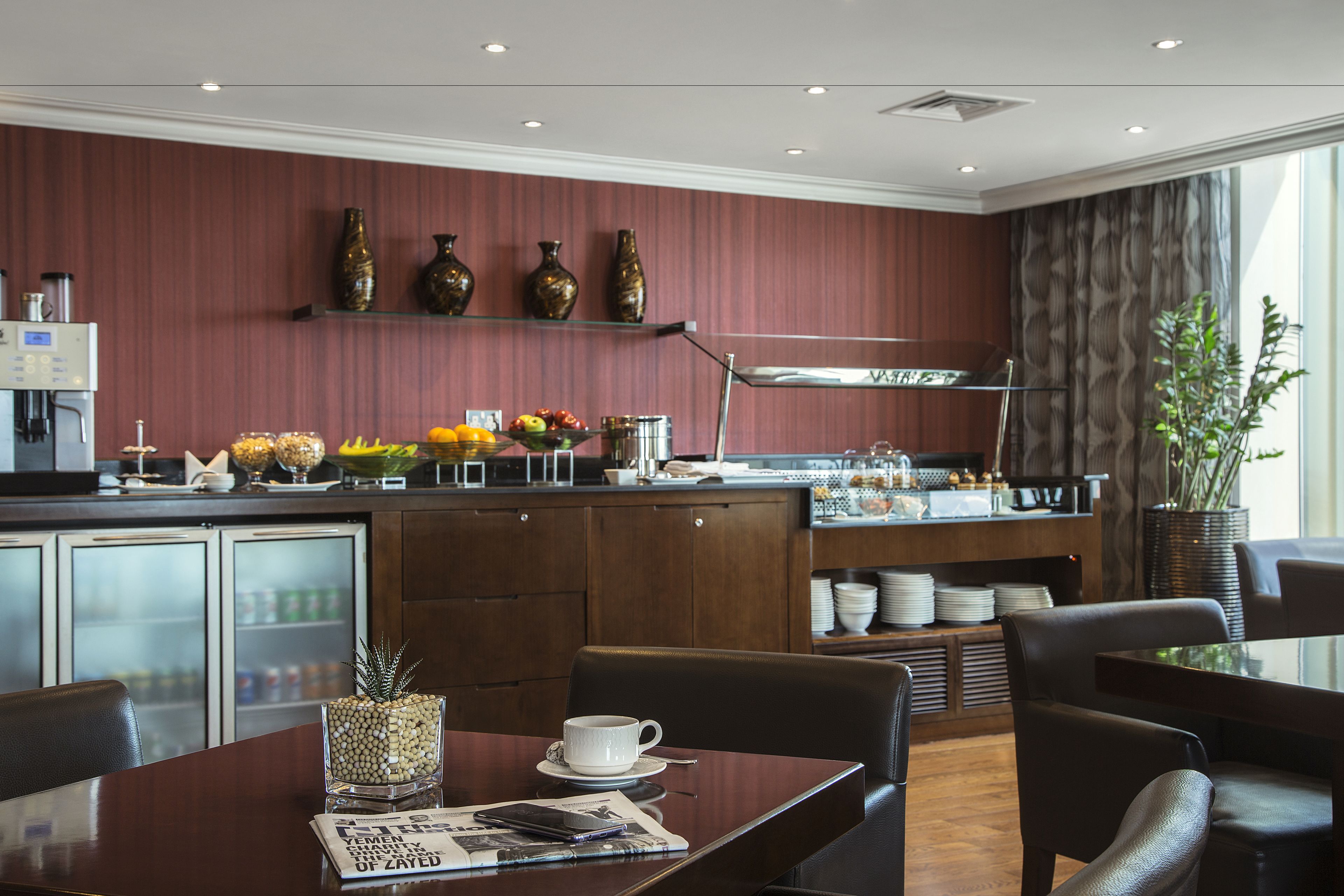 A modern dining area with a wooden table, chairs, a coffee station, and a kitchen in the background.