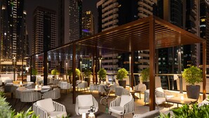 An outdoor seating area with white cushioned chairs and tables, surrounded by potted plants and a wooden pergola structure. - Towers Rotana (Dubai)