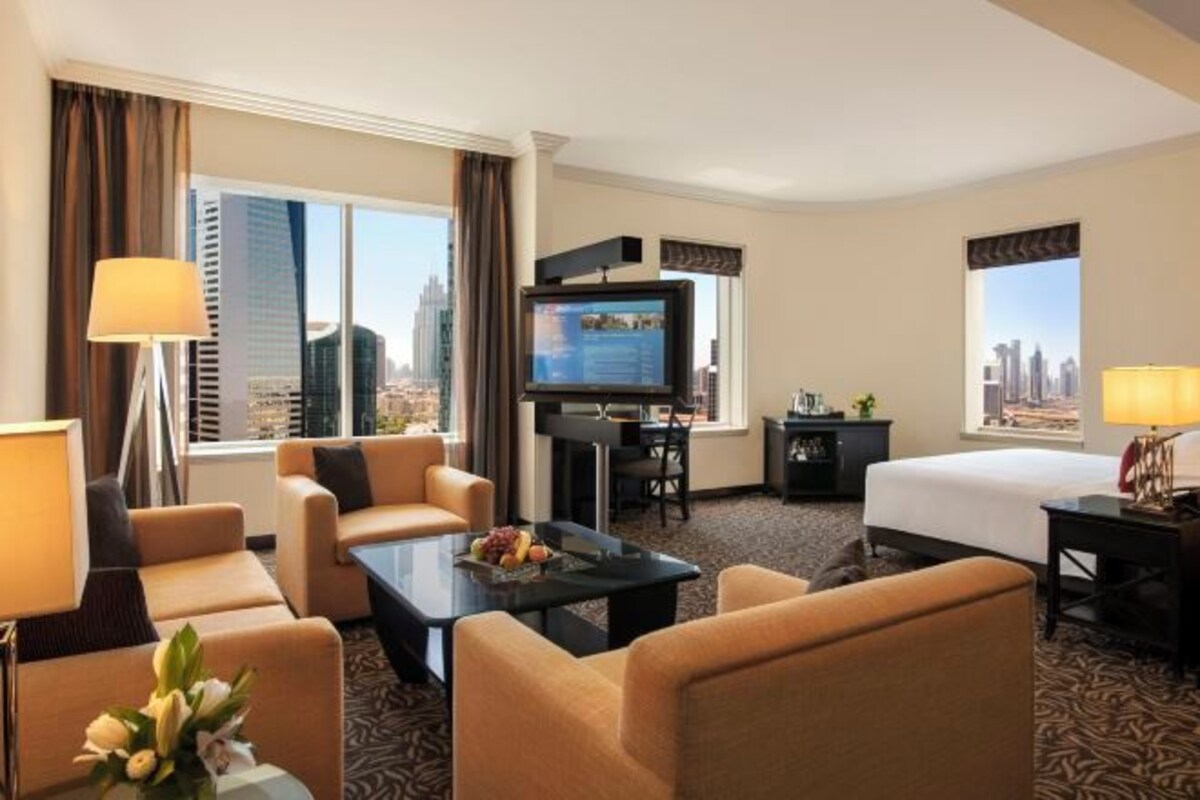 A hotel room with a bed, two armchairs, a coffee table, a TV, a desk, and a view of a cityscape.