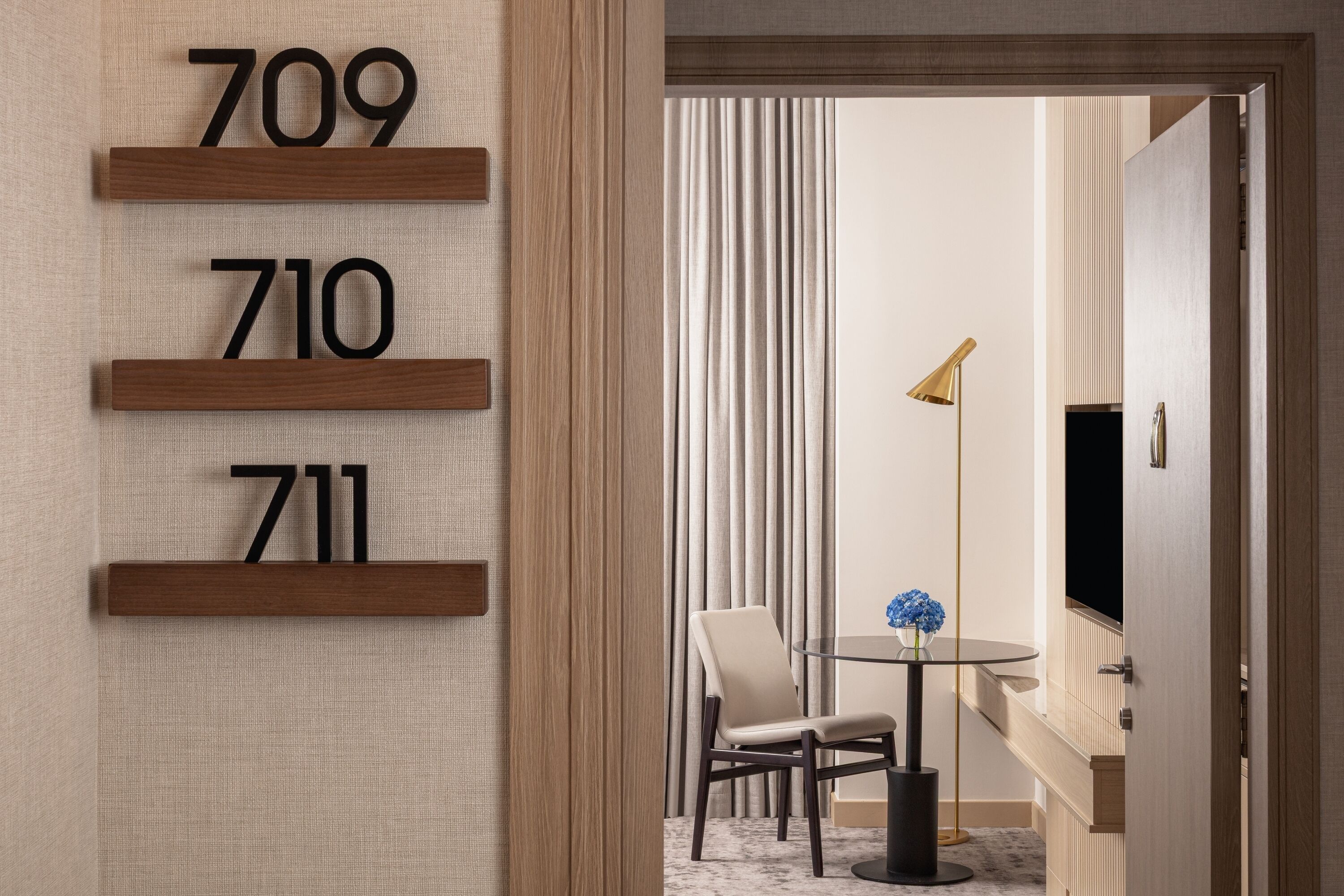 Habitación de hotel con un letrero de madera con un número, una mesa redonda con una silla, una lámpara de pie y un televisor en la pared.