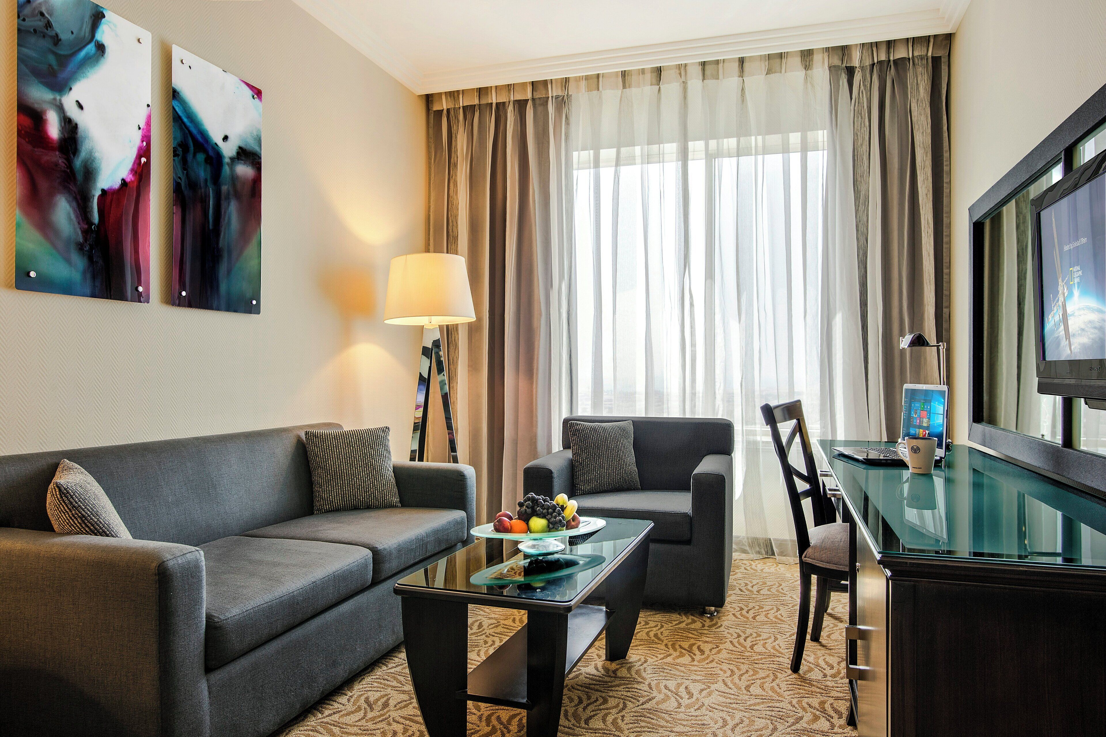 Una habitación de hotel moderna con sofá, sillón, una mesita de centro de vidrio y televisor sobre un mueble.