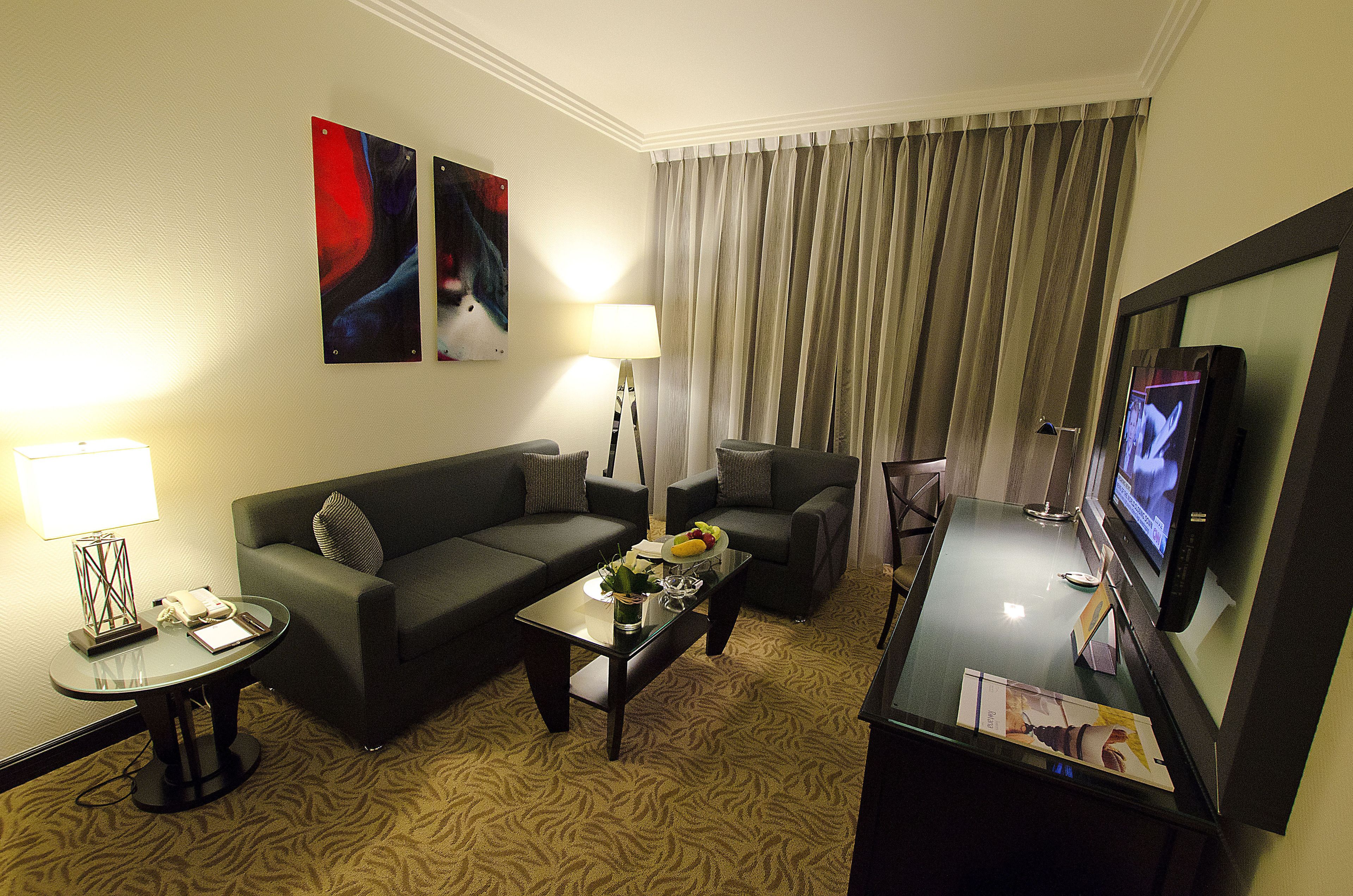 A hotel room with a sofa, armchairs, a coffee table, a TV, and a lamp.