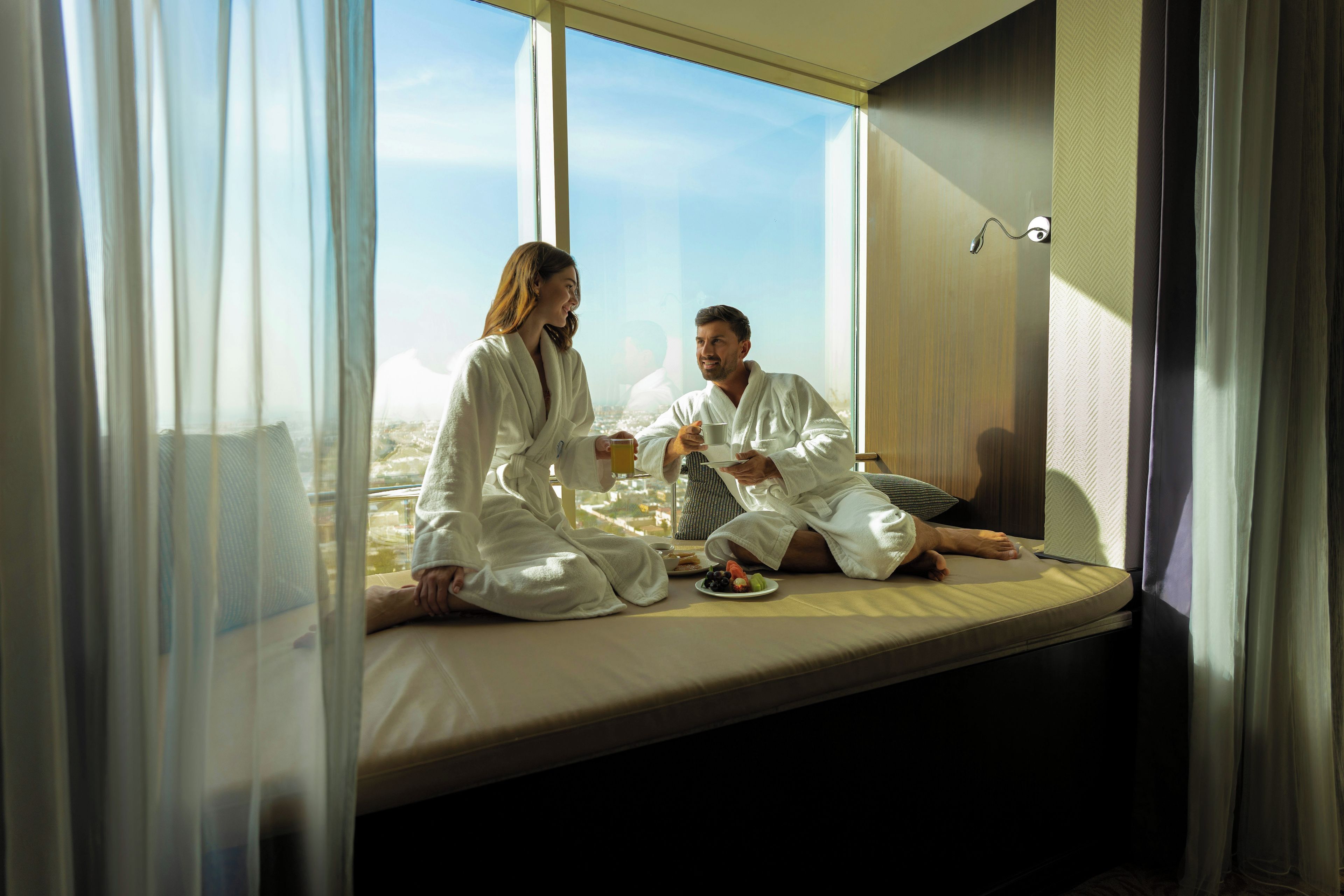 A couple in a hotel room with a large window, sitting on a bed with a view of the city.