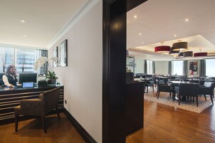 A hotel reception area with a receptionist, a computer, a potted plant, and a view of the dining area with tables and chairs.