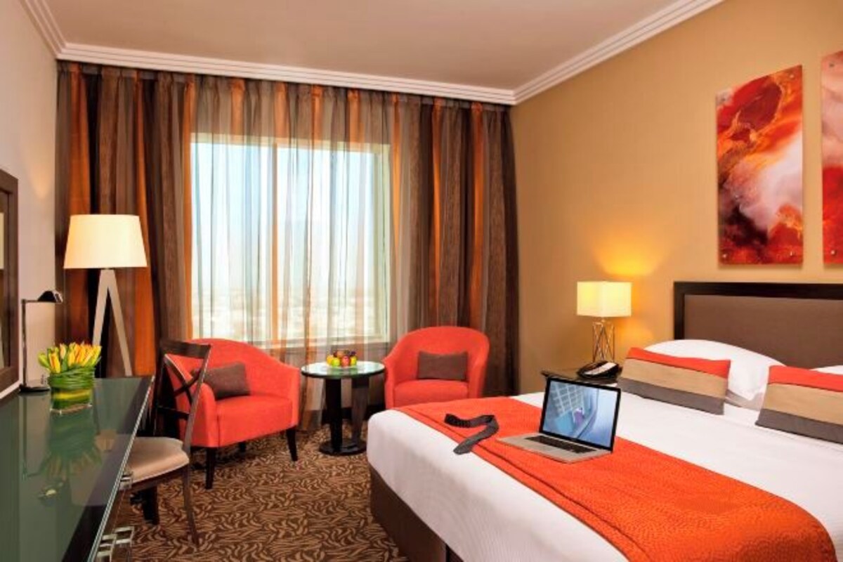A hotel room with a bed, two red chairs, a desk with a lamp, a laptop, and a view of the outdoors through curtains.