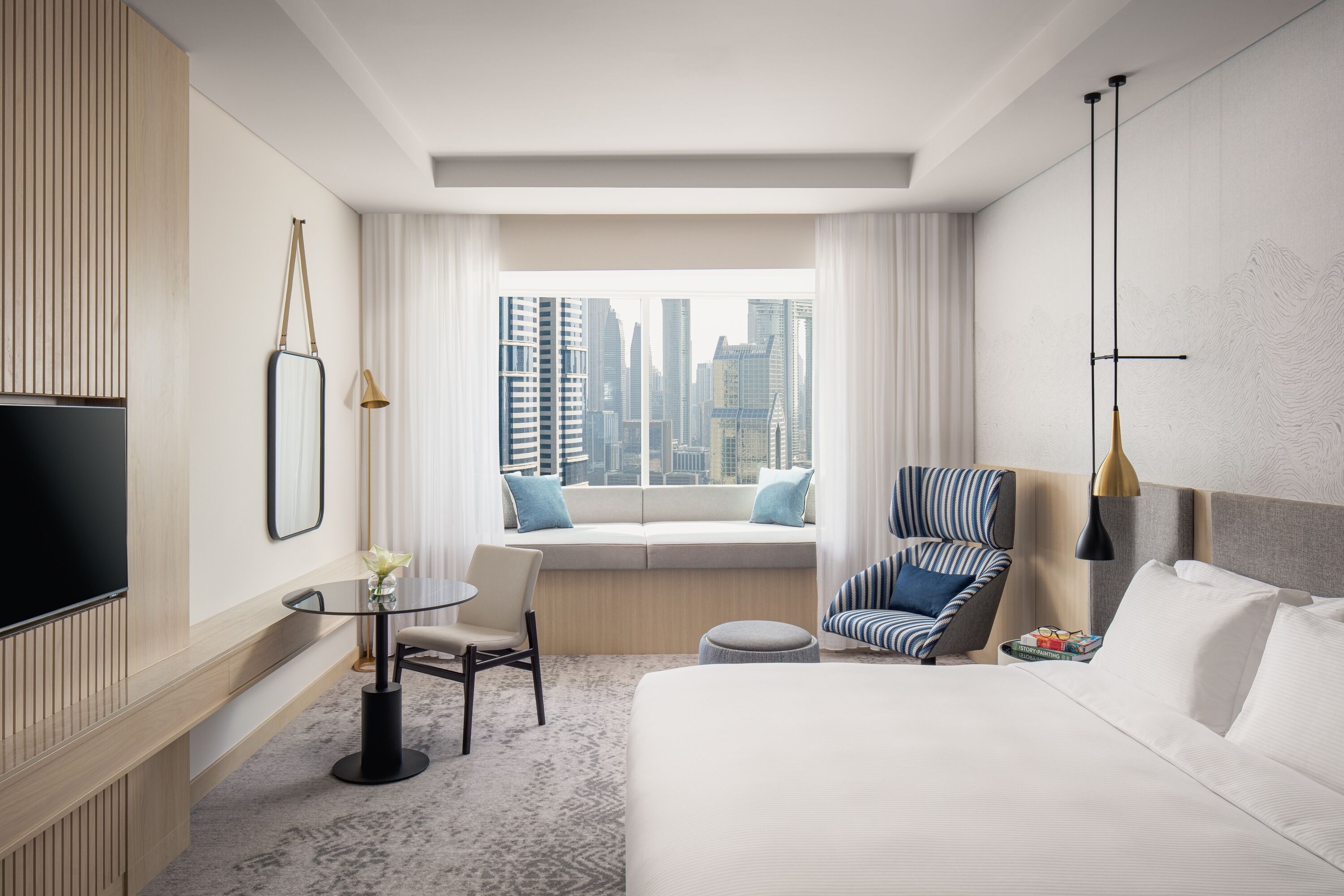 A modern hotel room with a large bed, a seating area with a sofa and armchairs, a small table with a chair, a TV, and a window with a city view.