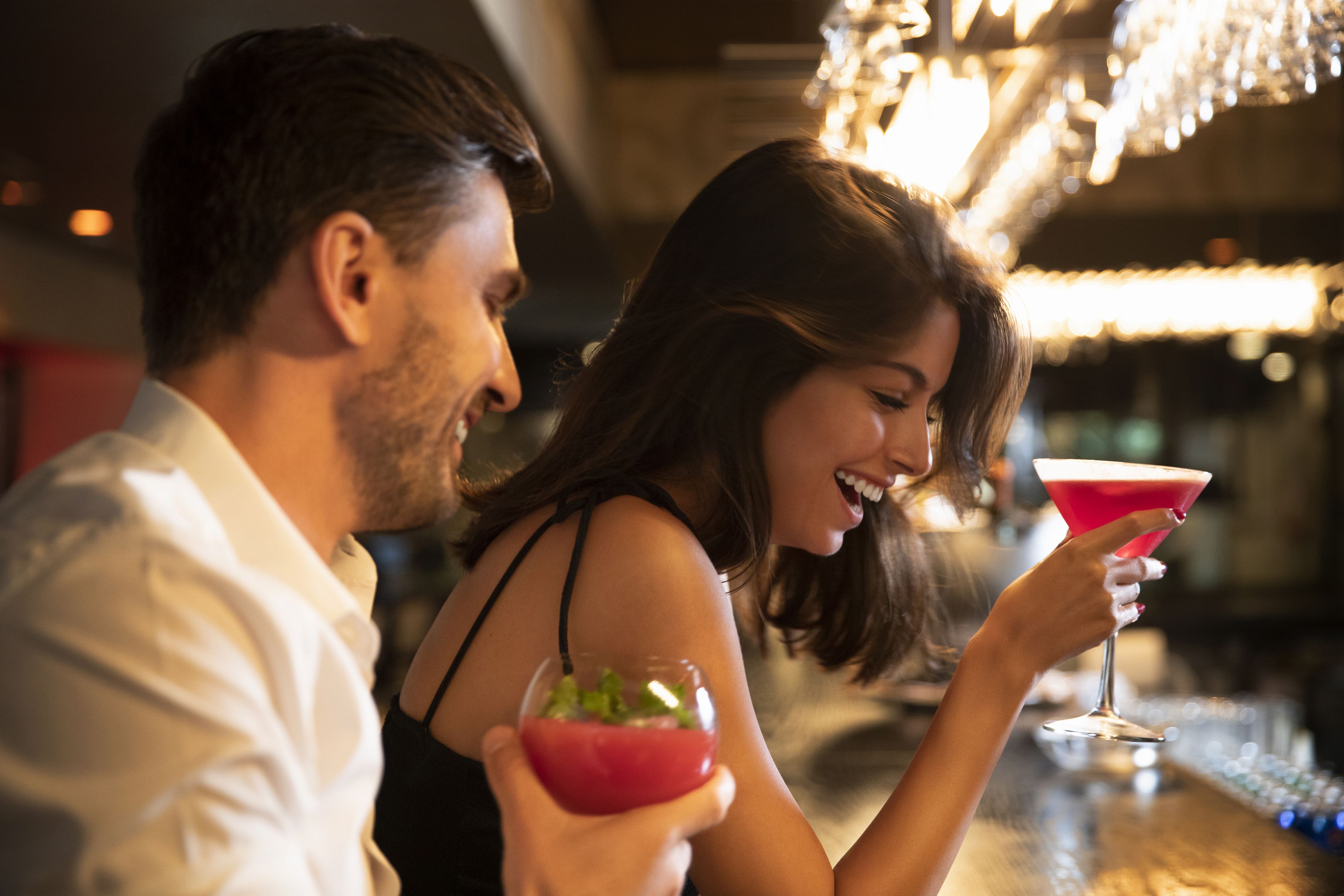 A couple seated at a bar with drinks, a chandelier in the background, and a blurred interior suggesting a cozy indoor setting.
