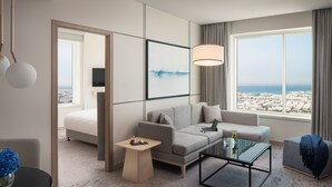 A modern hotel room with a sofa, a coffee table, a TV, and a large window with a city view. - Towers Rotana (Dubai)