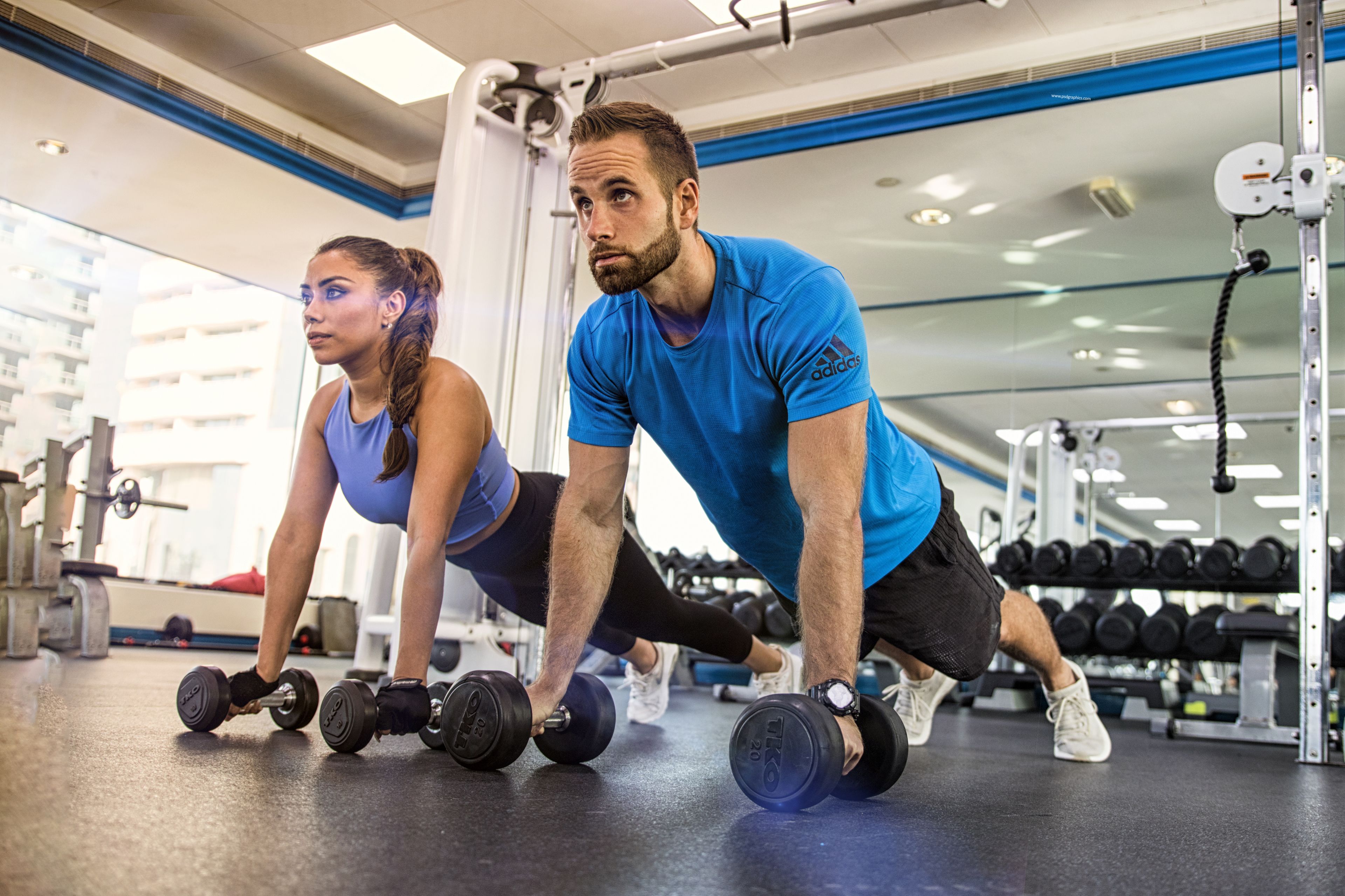 Two people doing a workout with dumbbells in a gym with various equipment.