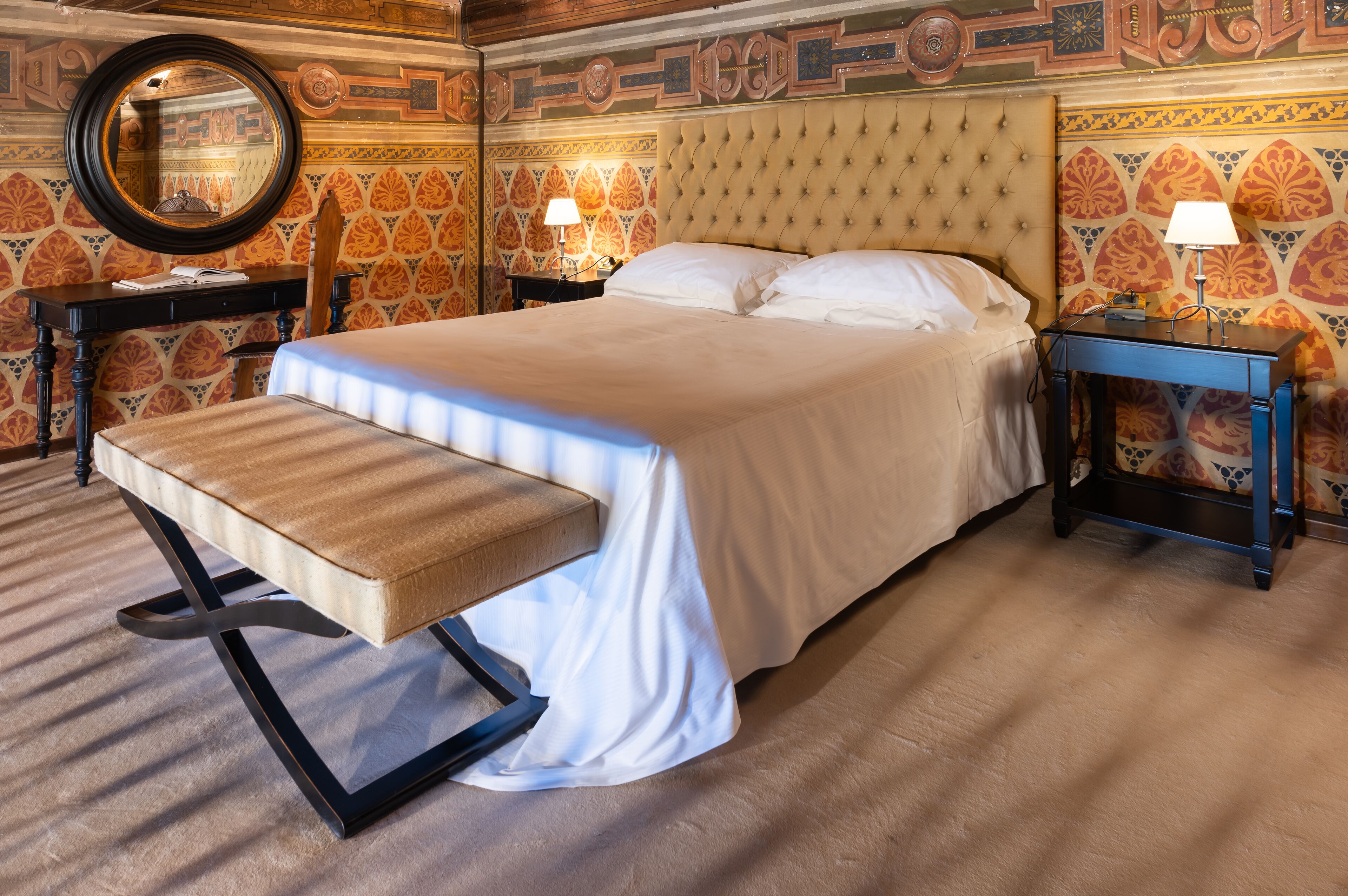 A neatly made bed with a bench, two bedside tables, a round mirror, and patterned wallpaper.