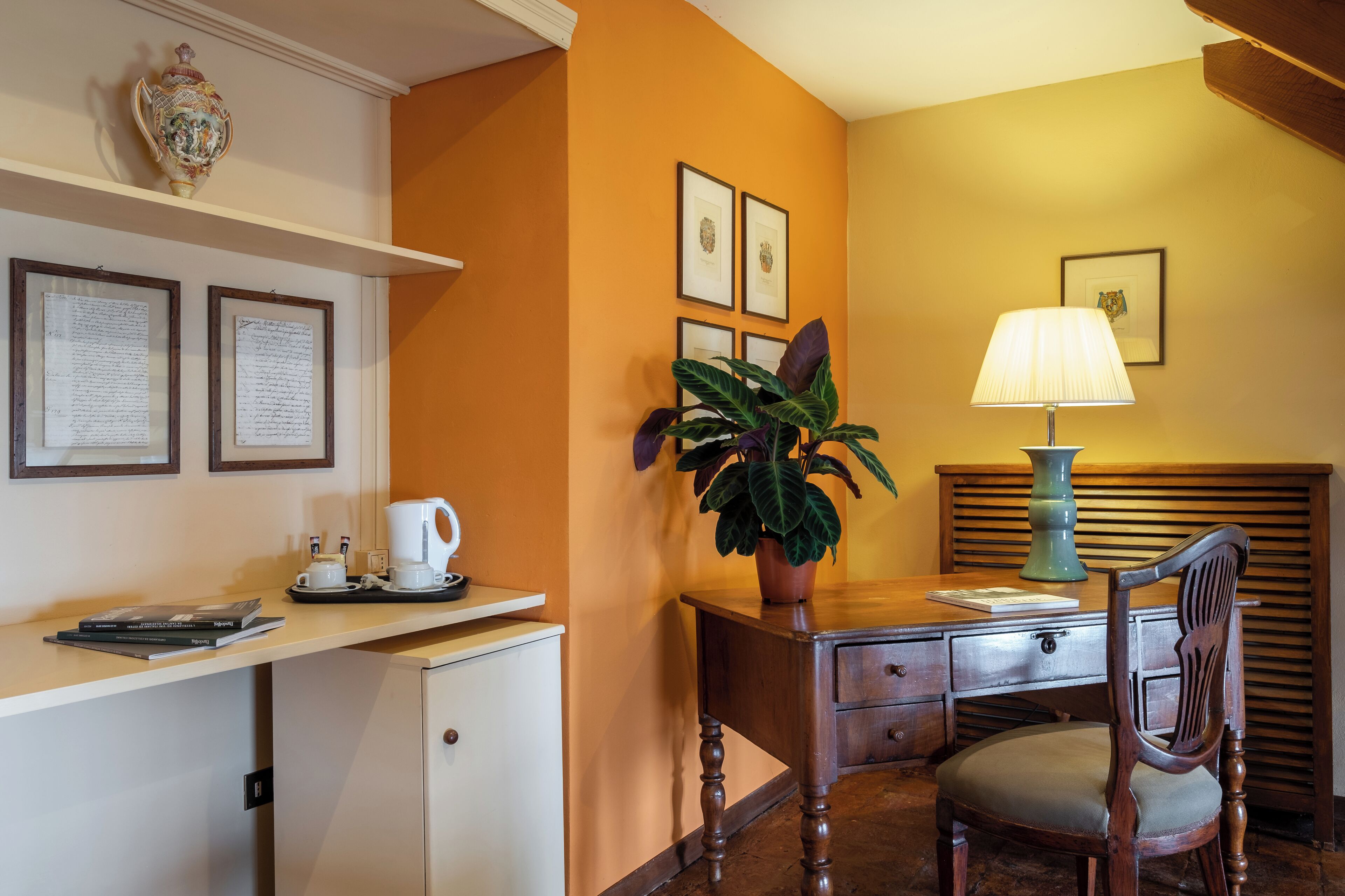 A hotel room with a desk, a chair, a lamp, a potted plant, and framed pictures on the wall.