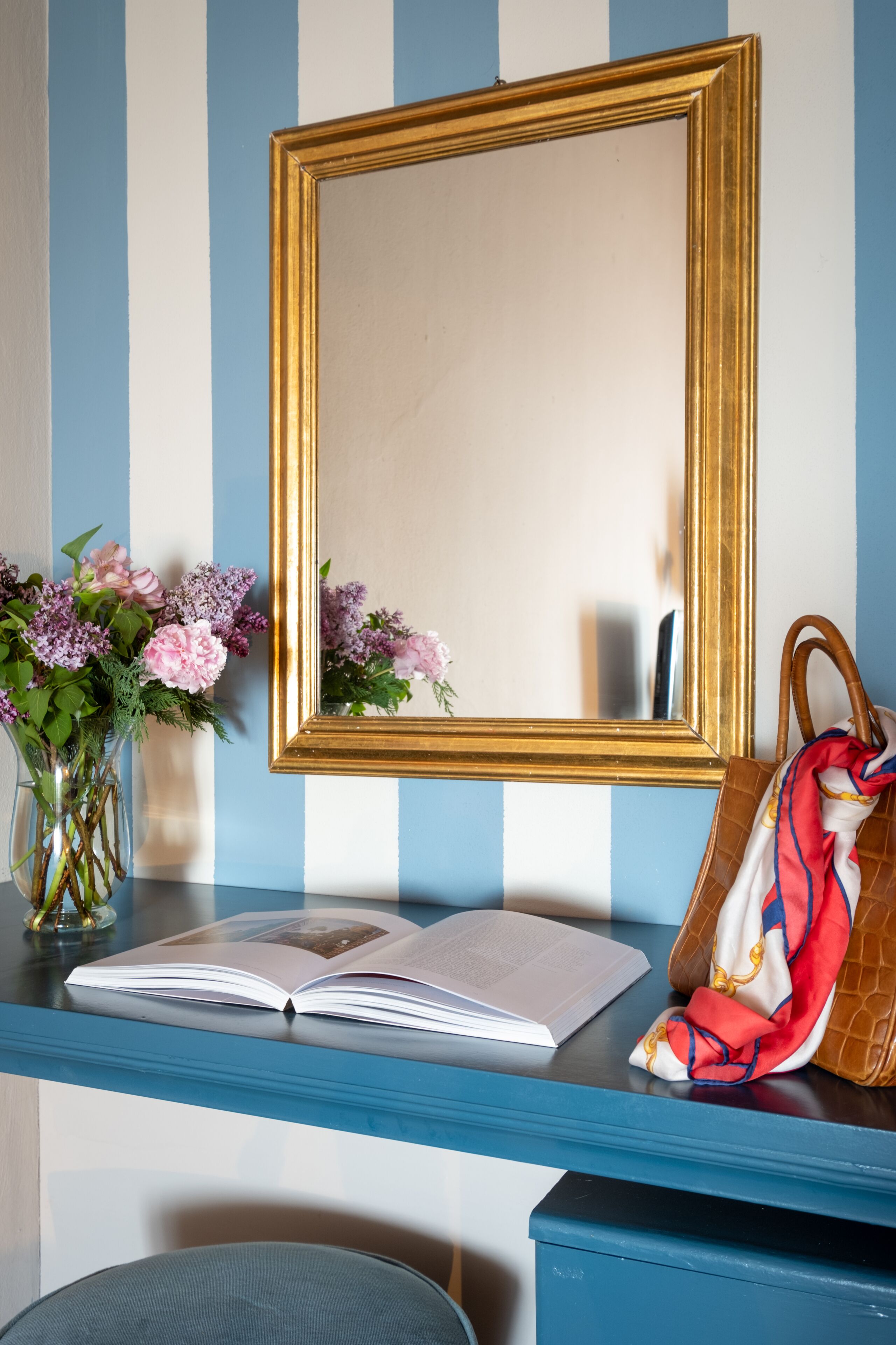 Eine blaue und weiße Streifenwand, ein goldrahmiger Spiegel, eine Vase mit Blumen, ein Buch und eine Handtasche auf einem blauen Schreibtisch.