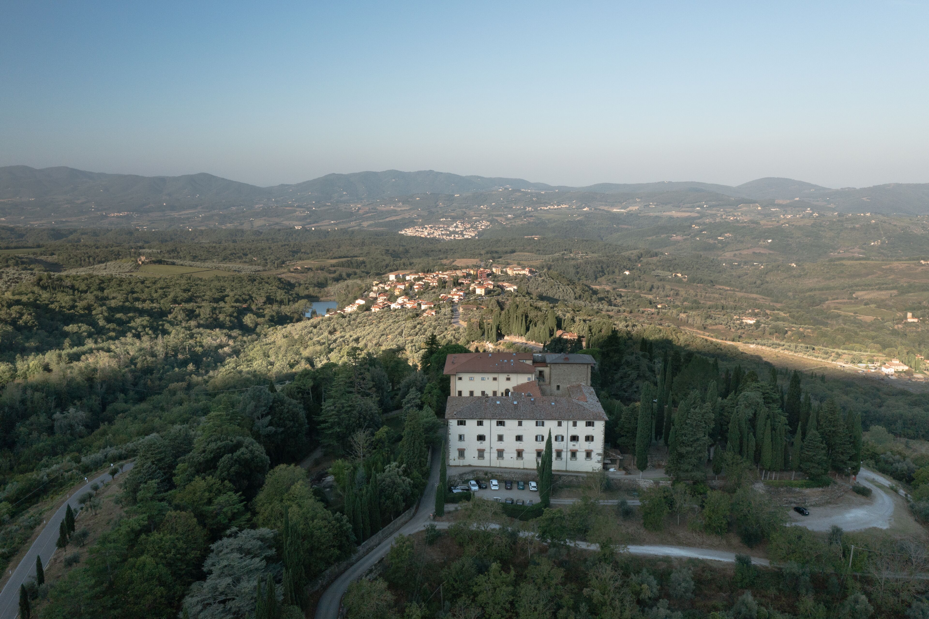 Un edificio grande con varios pisos, rodeado de un bosque denso y colinas al fondo.