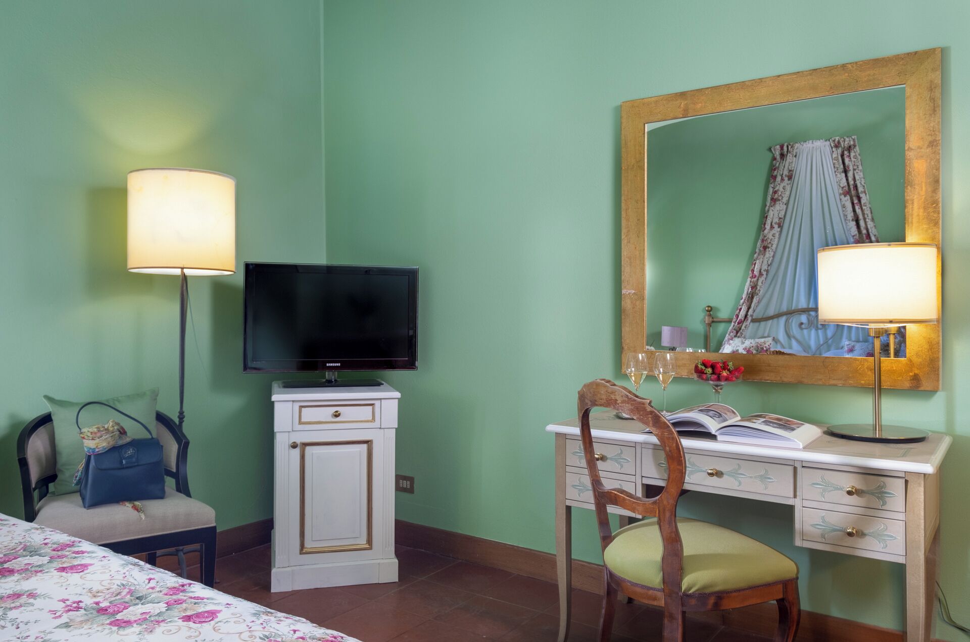 A hotel room with a bed, a desk with a chair, a television, a mirror, and a lamp.