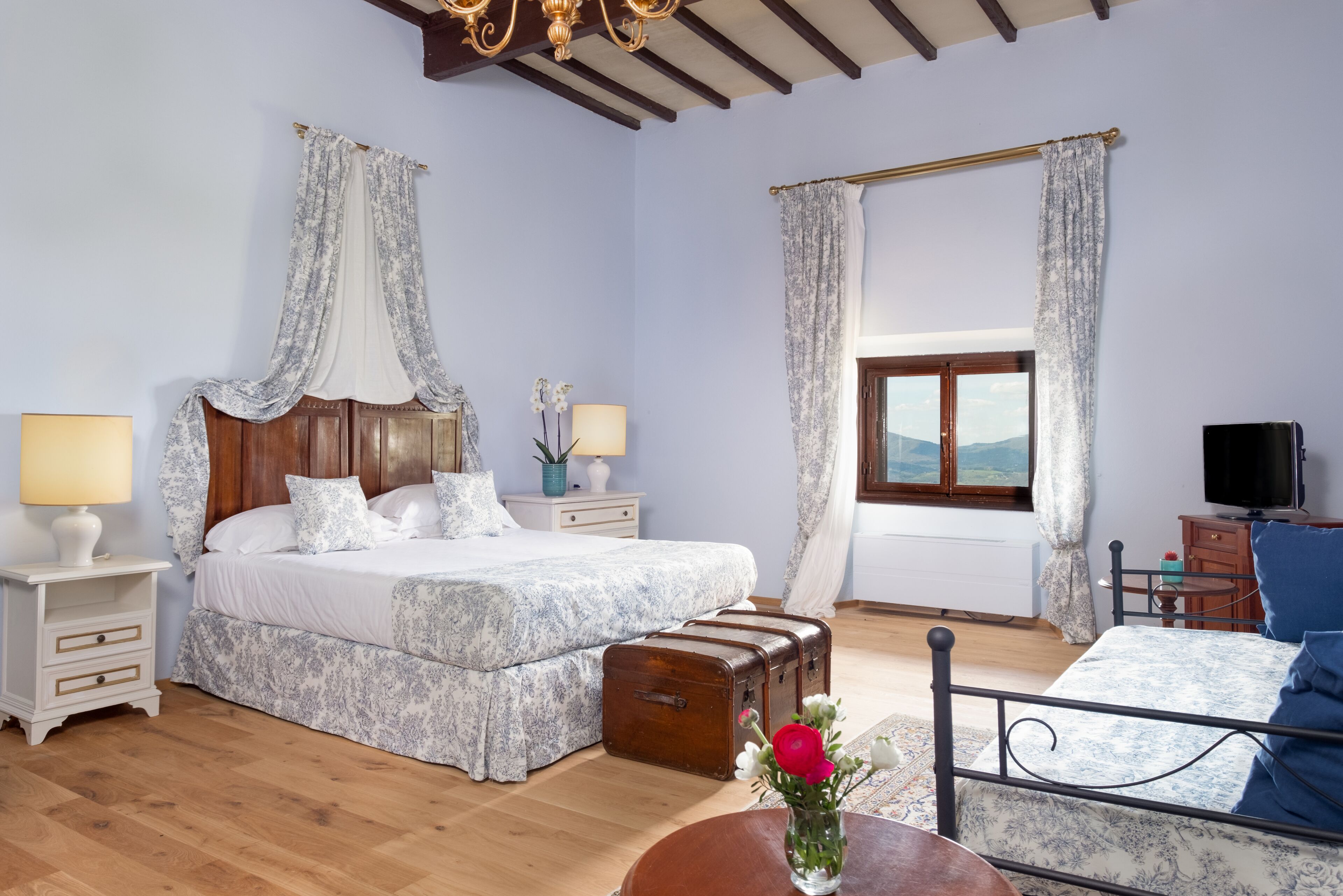 A bedroom with a large bed, a wooden headboard, two bedside tables with lamps, a suitcase, a small table with a vase, and a TV.