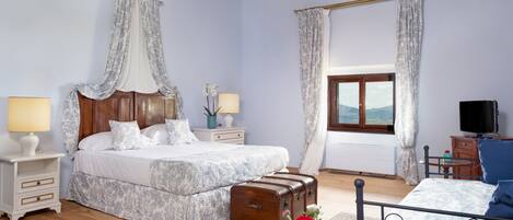 A bedroom with a large bed, a wooden headboard, two bedside tables with lamps, a suitcase, a small table with a vase, and a TV.