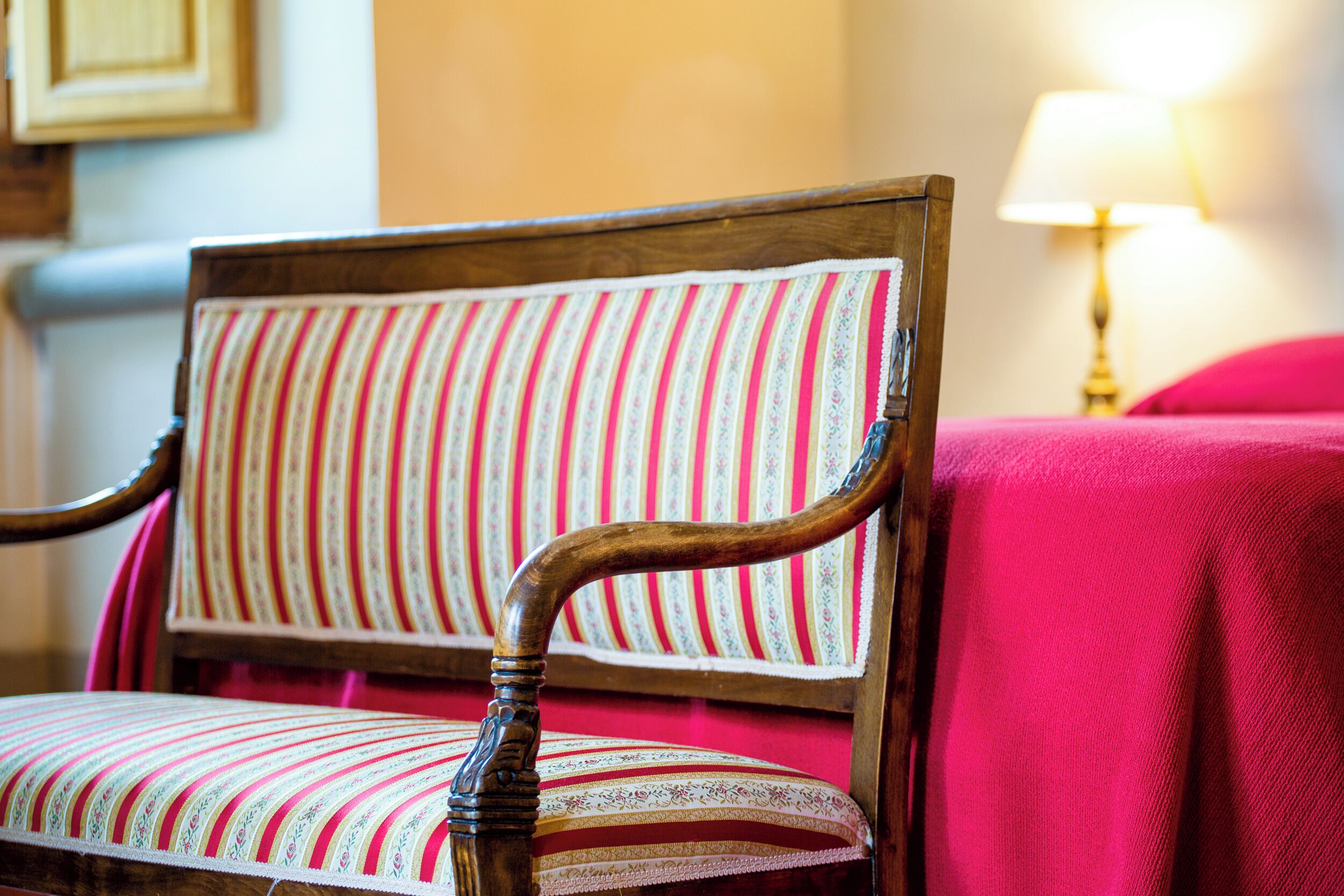 A wooden chair with a striped cushion in a room with a red bedspread and a lamp in the background.