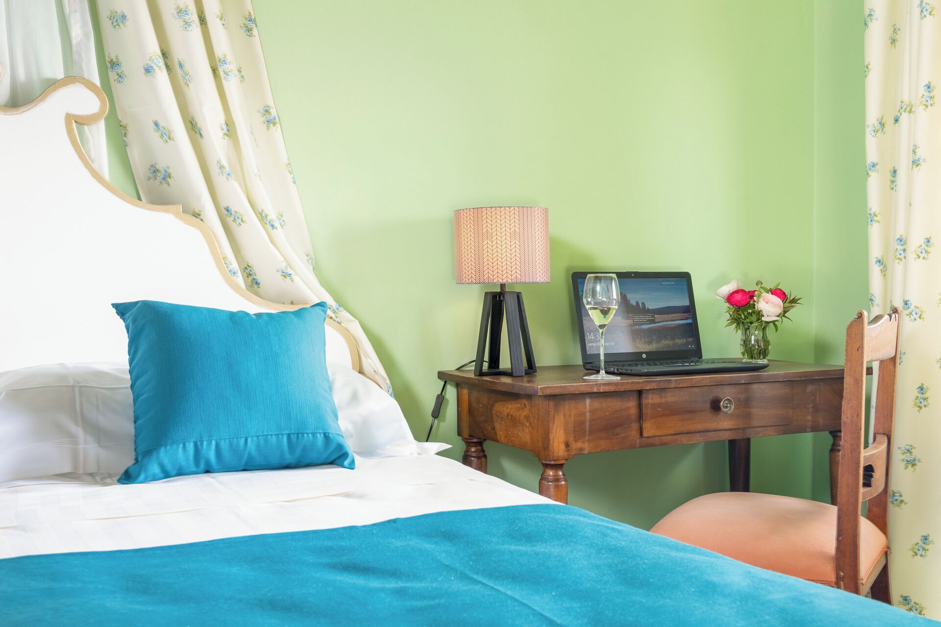 Una cama bien hecha con una almohada azul, una mesita de noche de madera con una laptop, una copa de vino y un florero con flores.