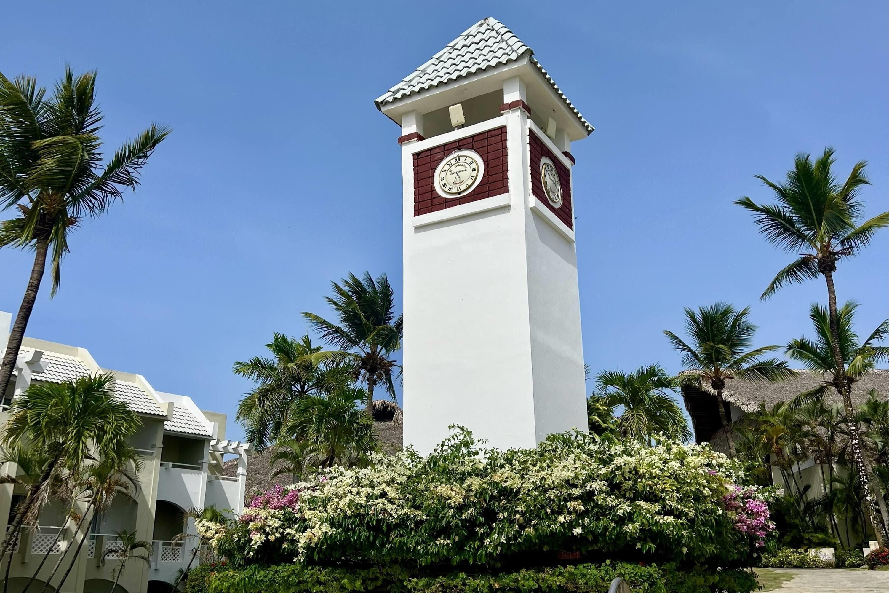 A clock tower with a white and red facade, surrounded by palm trees and flowering plants.
