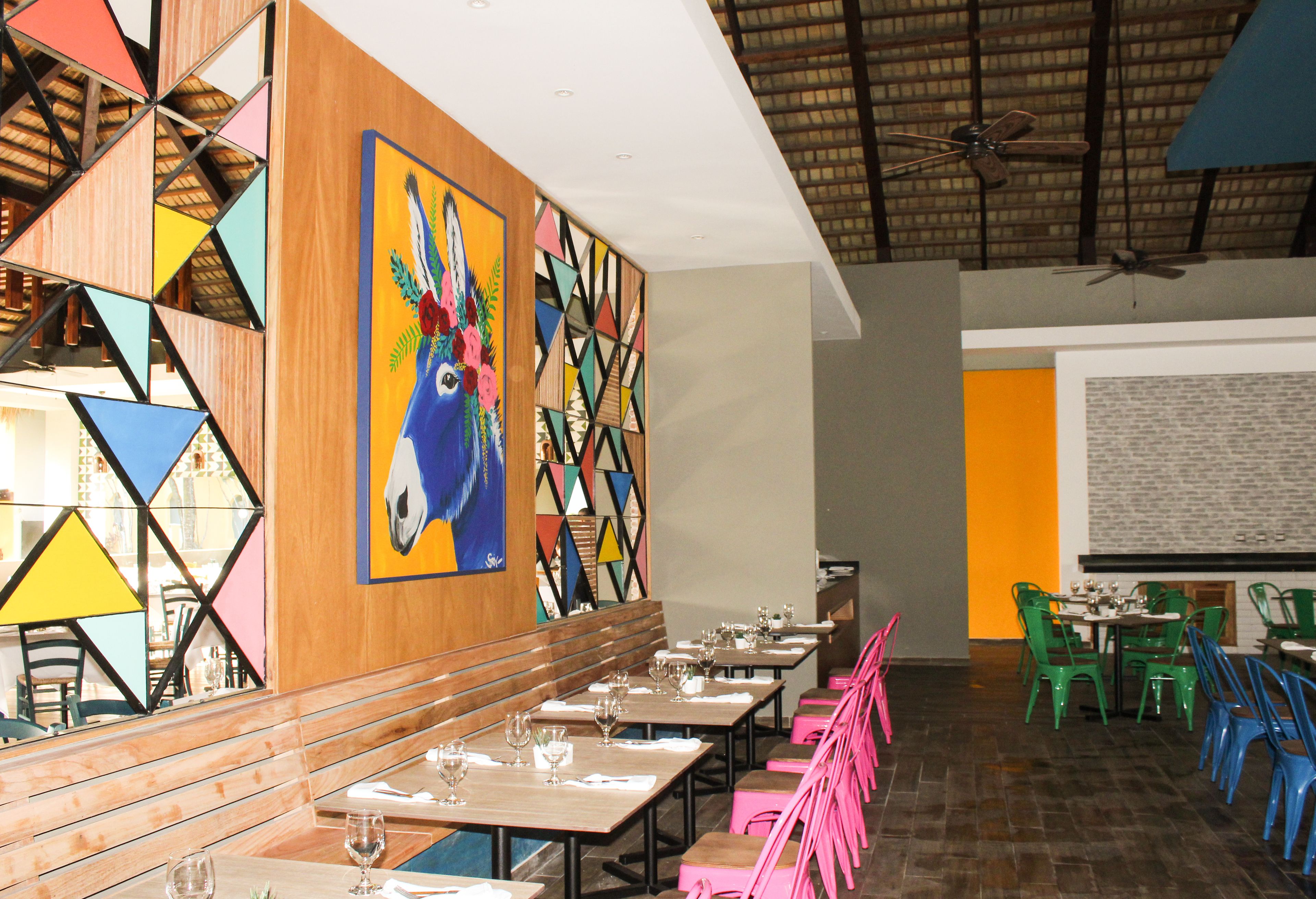 Un restaurante con largos bancos y mesas de madera, arte mural geométrico colorido y un ventilador de techo.