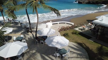 A beachside patio with tables and chairs under umbrellas, overlooking the ocean.