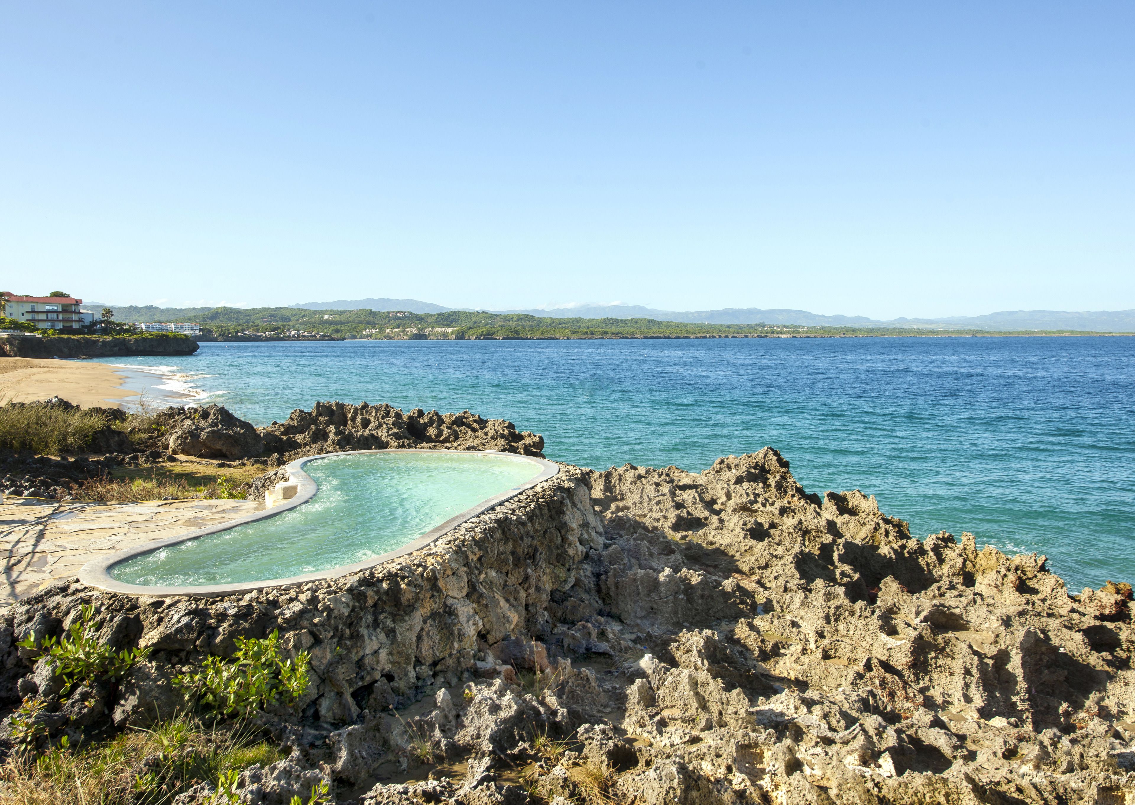 A rocky coastline with a clear blue sea, a swimming pool, and a building in the background.