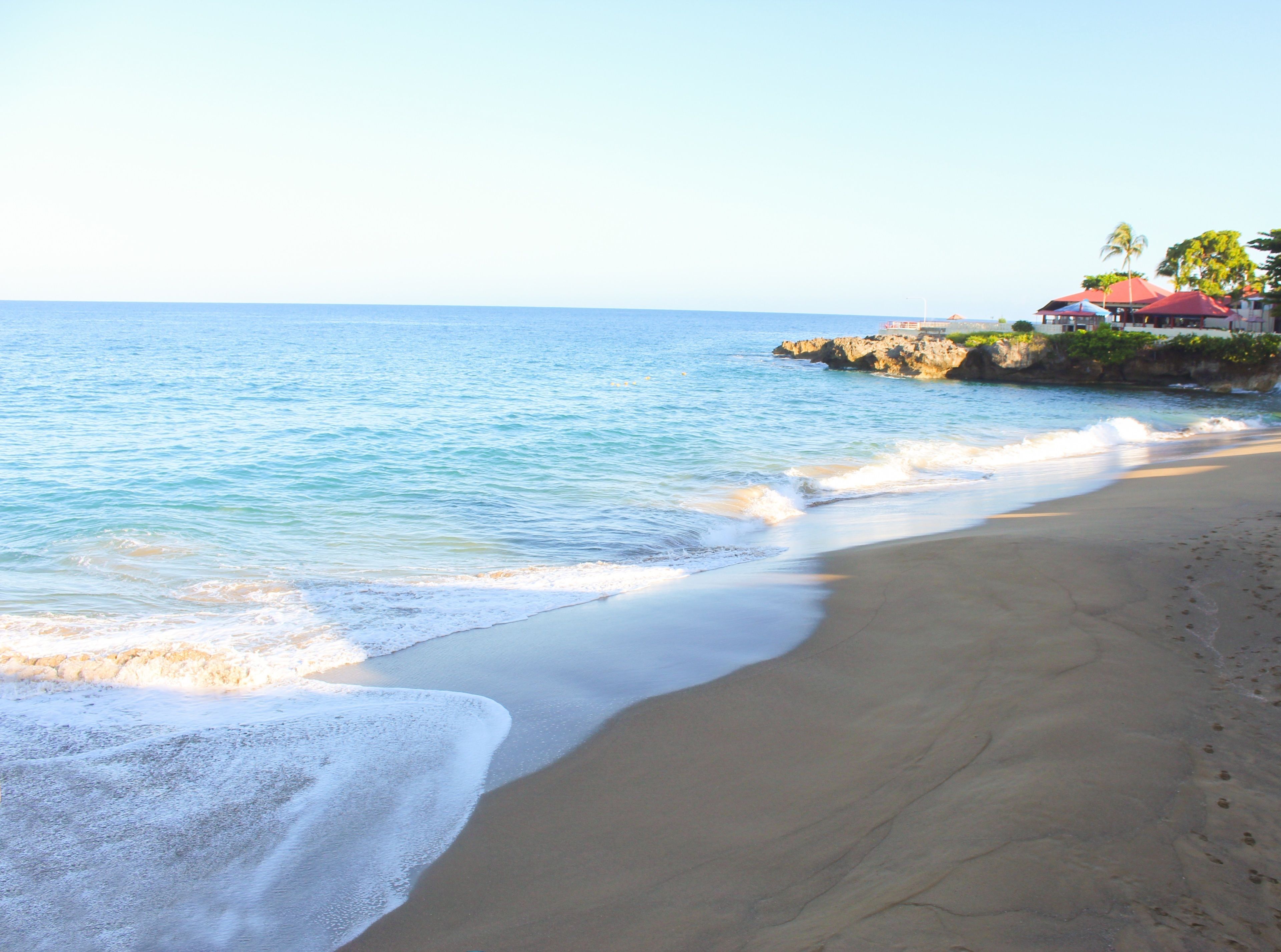 A beach with clear blue water, a sandy shore, and a building with a red roof near the water's edge.