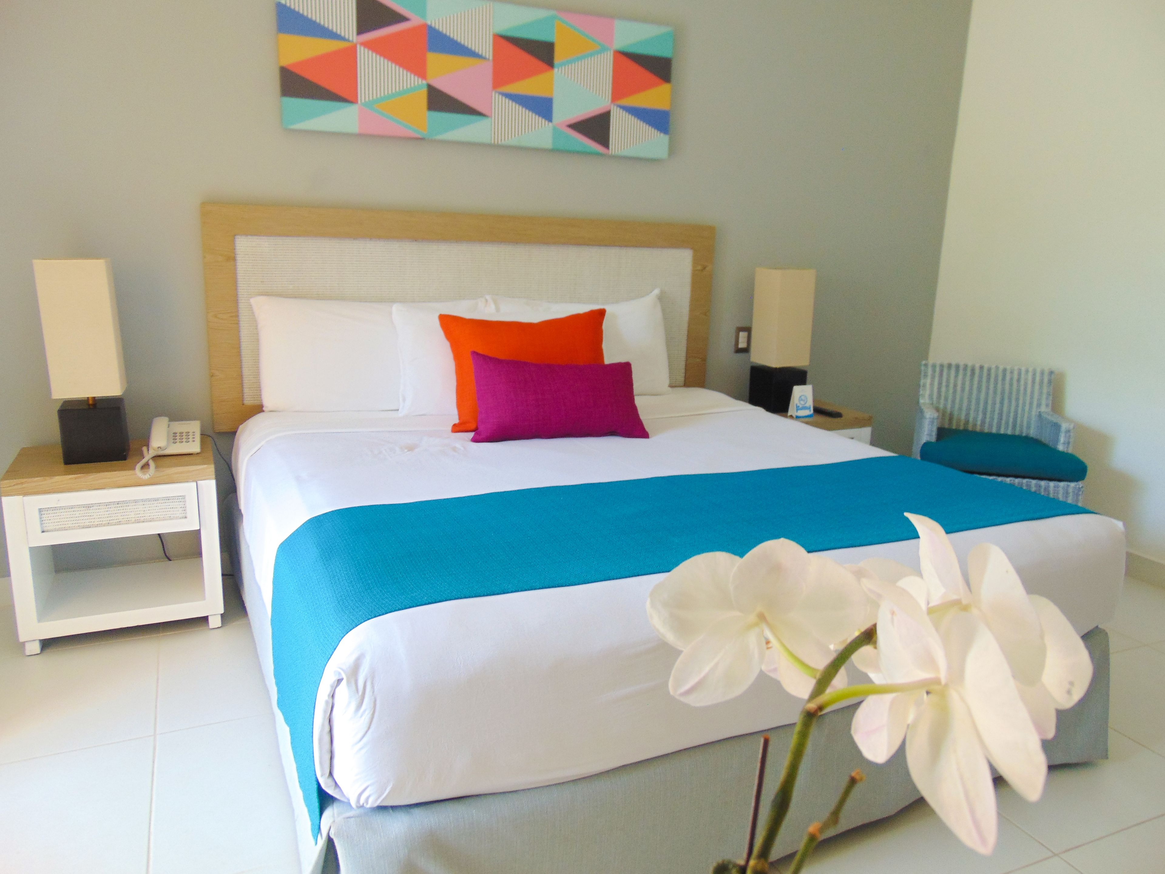 Een hotelkamer met een groot bed, twee nachtkastjes, een stoel en een kleurrijk geometrisch kunstwerk aan de muur.