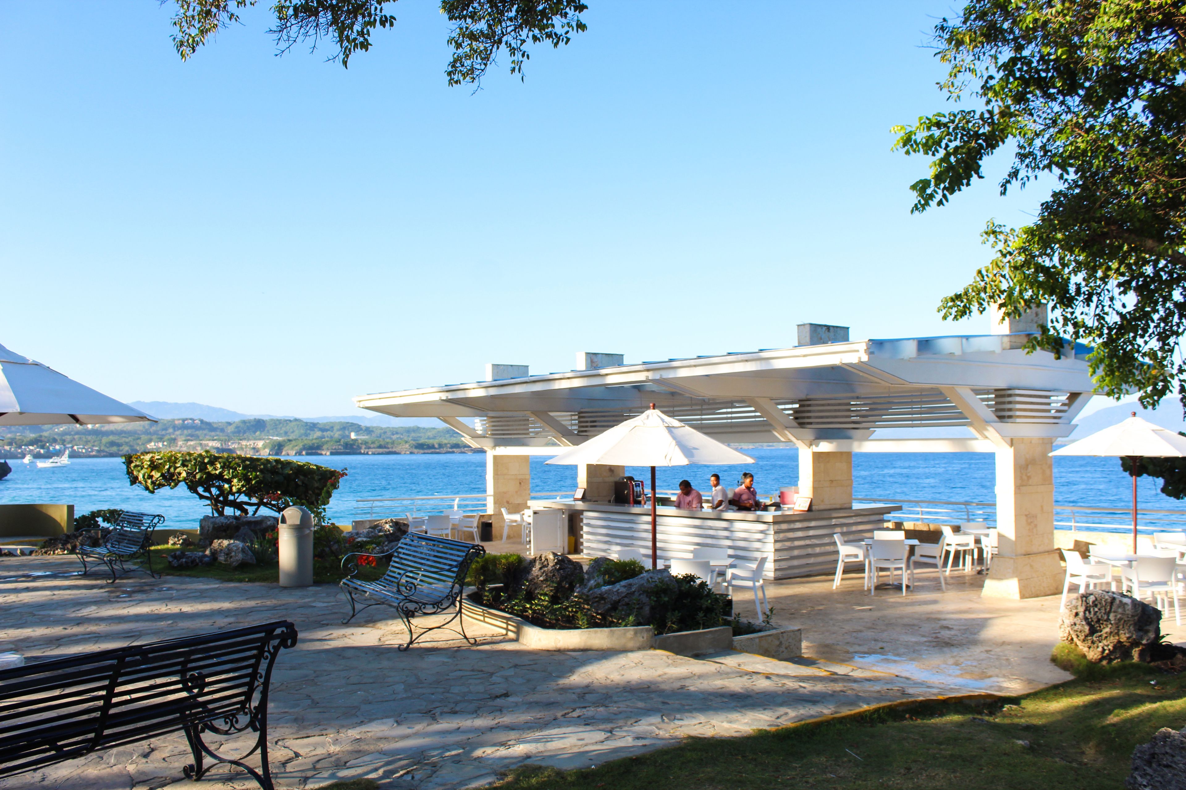 An outdoor seating area with stone flooring, white tables, and benches, overlooking a body of water.