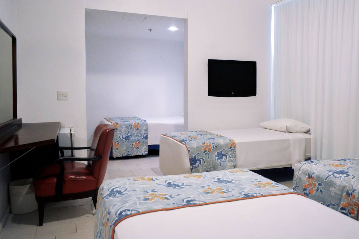 A hotel room with a desk, two beds with floral bedspreads, a TV mounted on the wall, and a window with curtains.