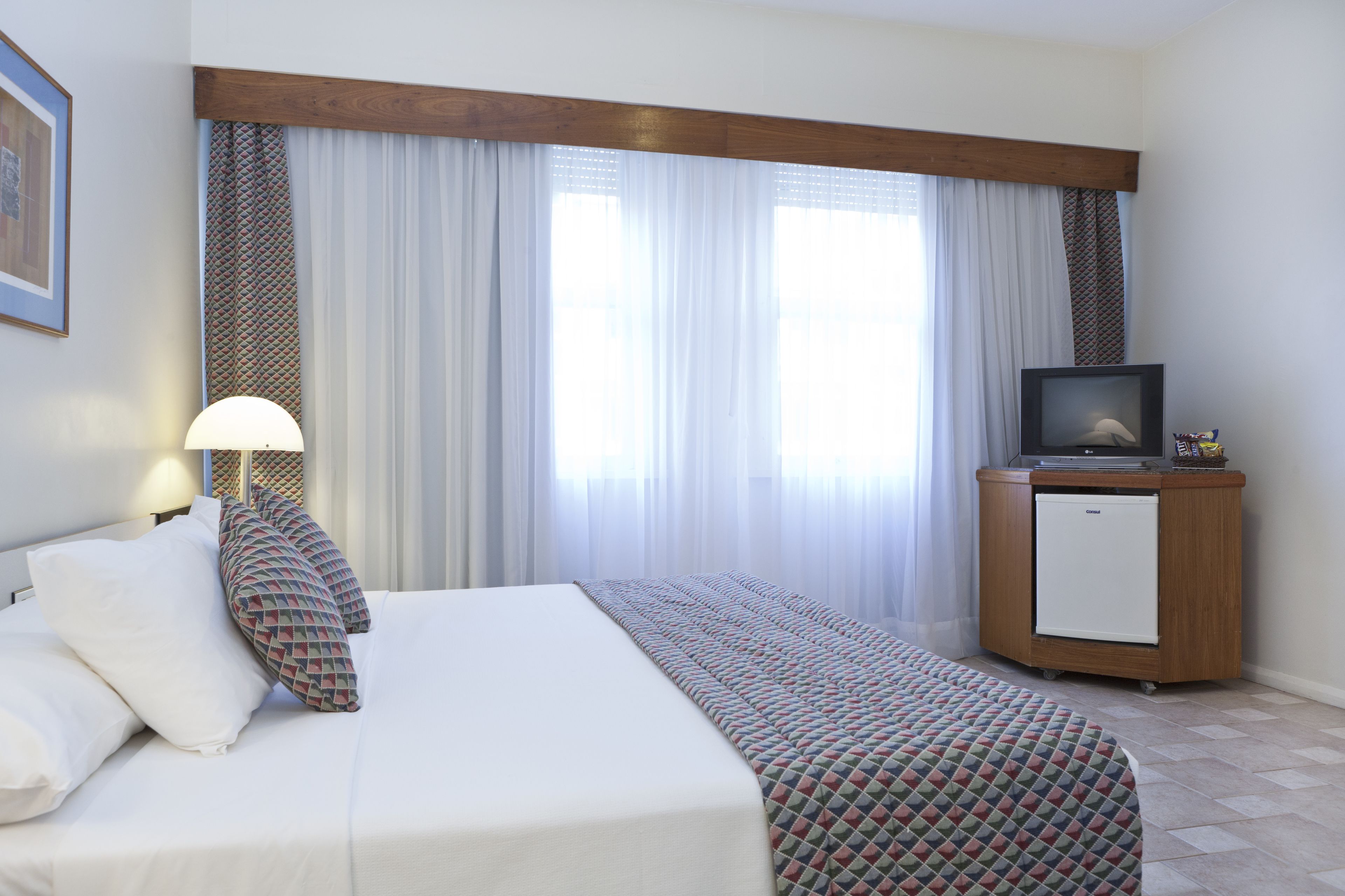 A hotel room with a bed, a TV, a small fridge, and a window with curtains.