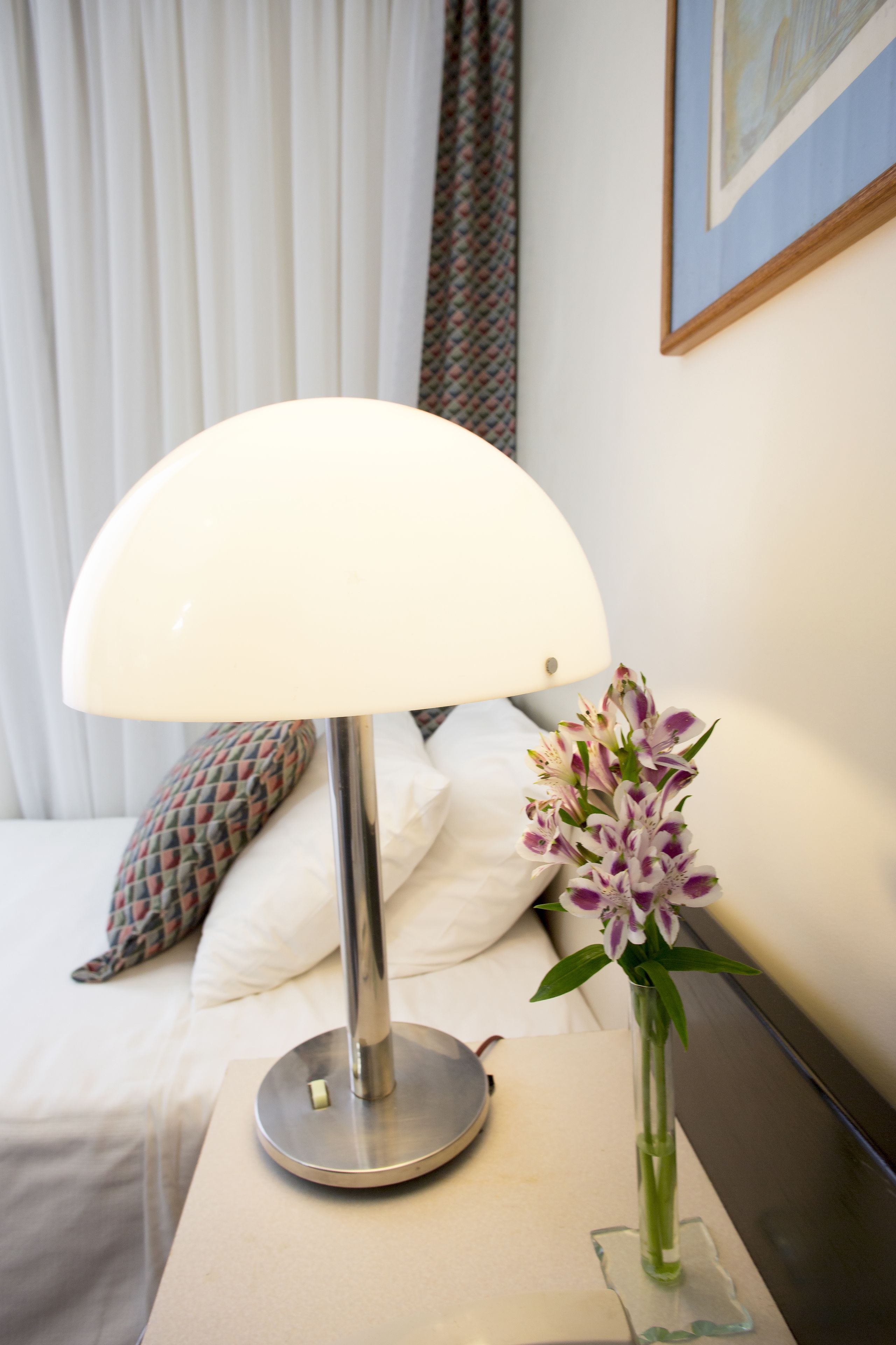 A hotel room with a bed, a bedside table with a lamp, and a vase with flowers.