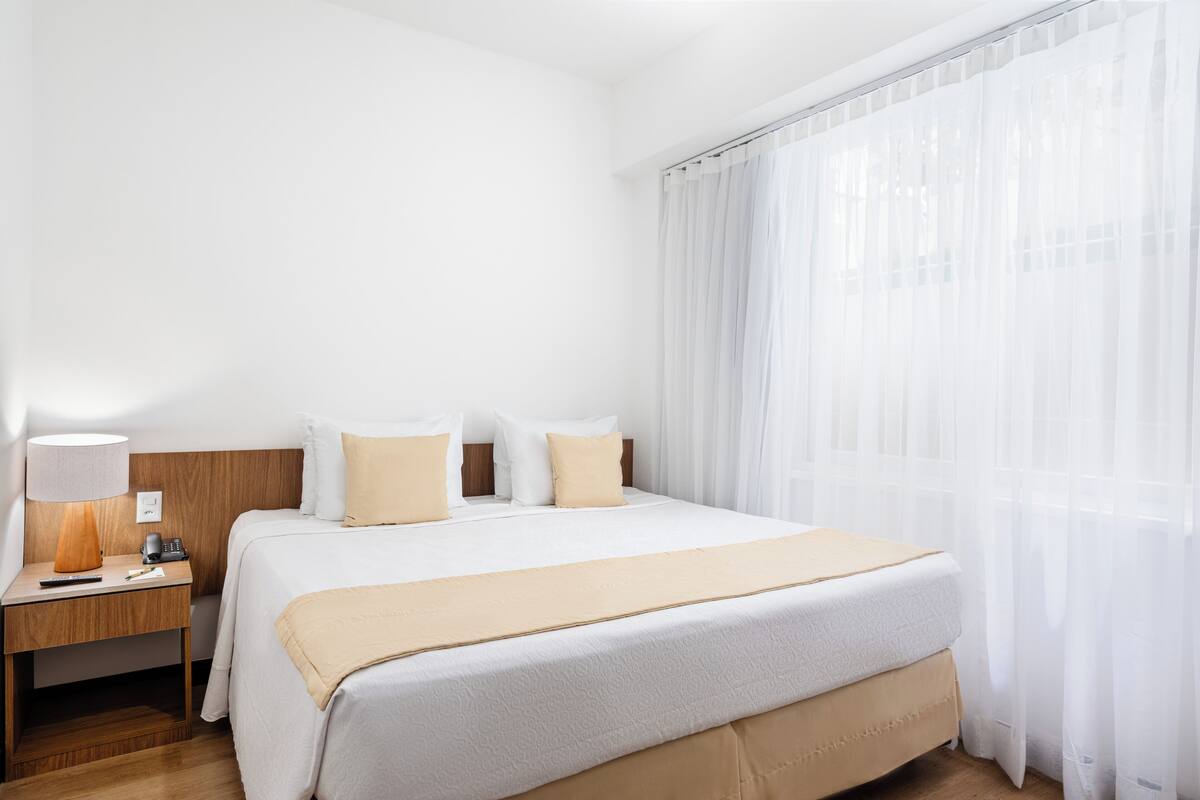 A hotel room with a large bed, a bedside table with a lamp, and sheer curtains.