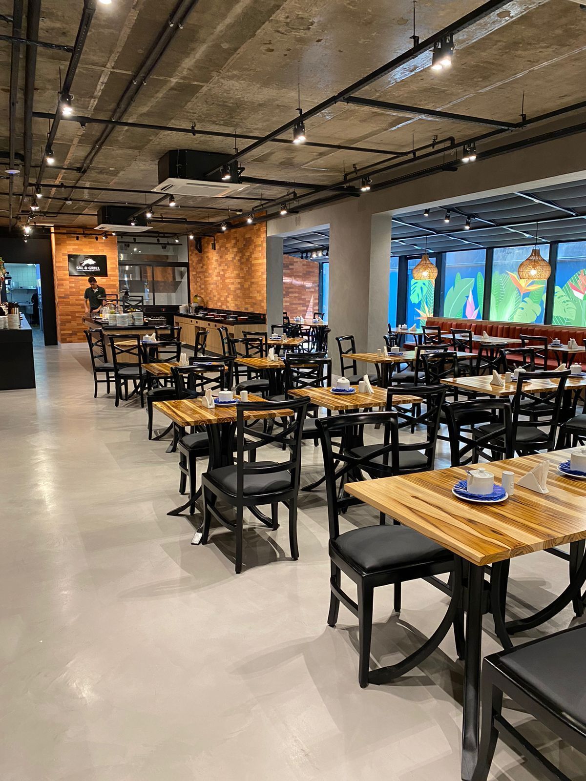 A modern restaurant interior with wooden tables and black chairs, a kitchen area, and a large window with a colorful mural.