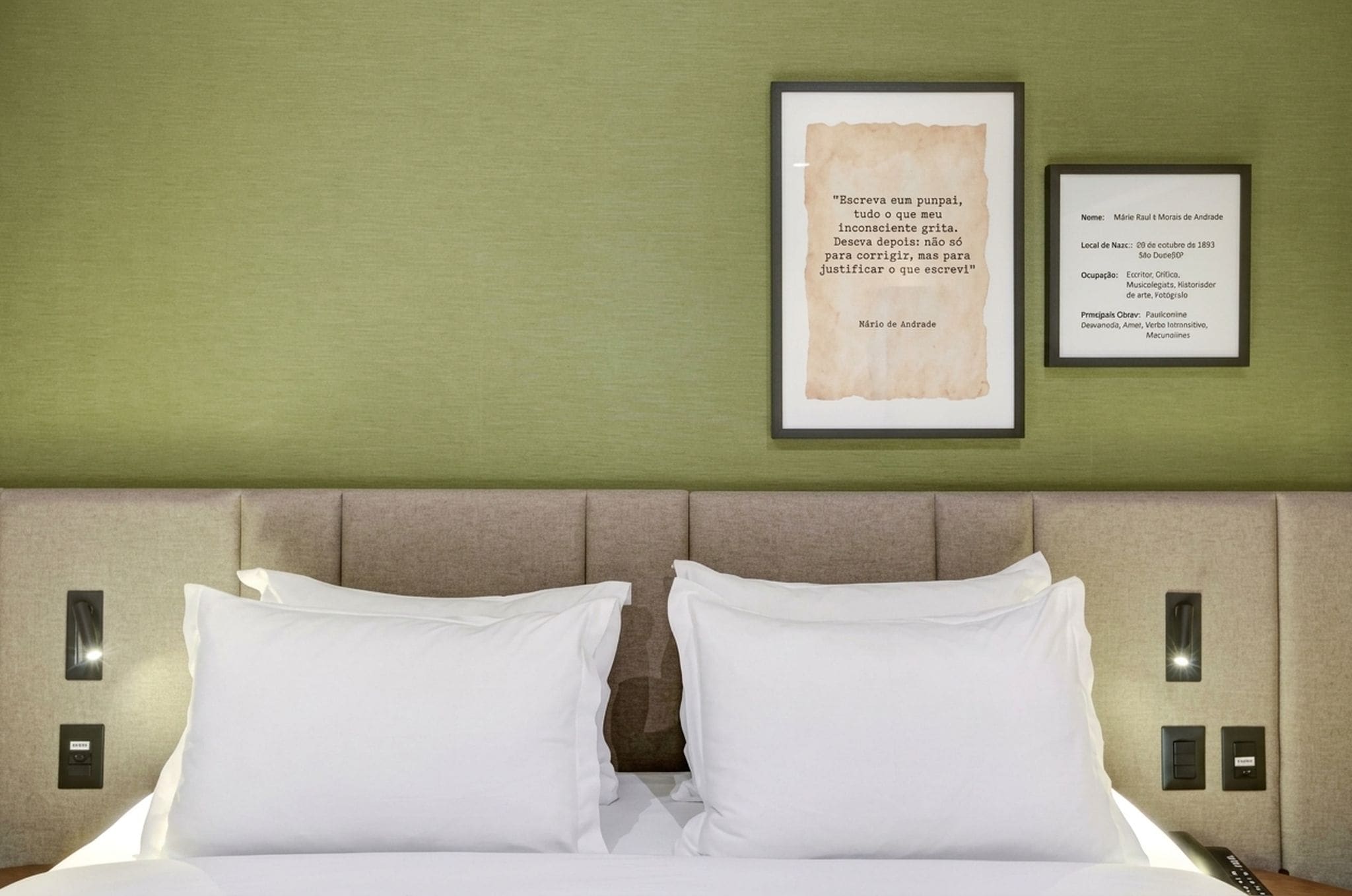 A hotel room with a bed, two pillows, a framed quote on the wall, and a framed certificate.