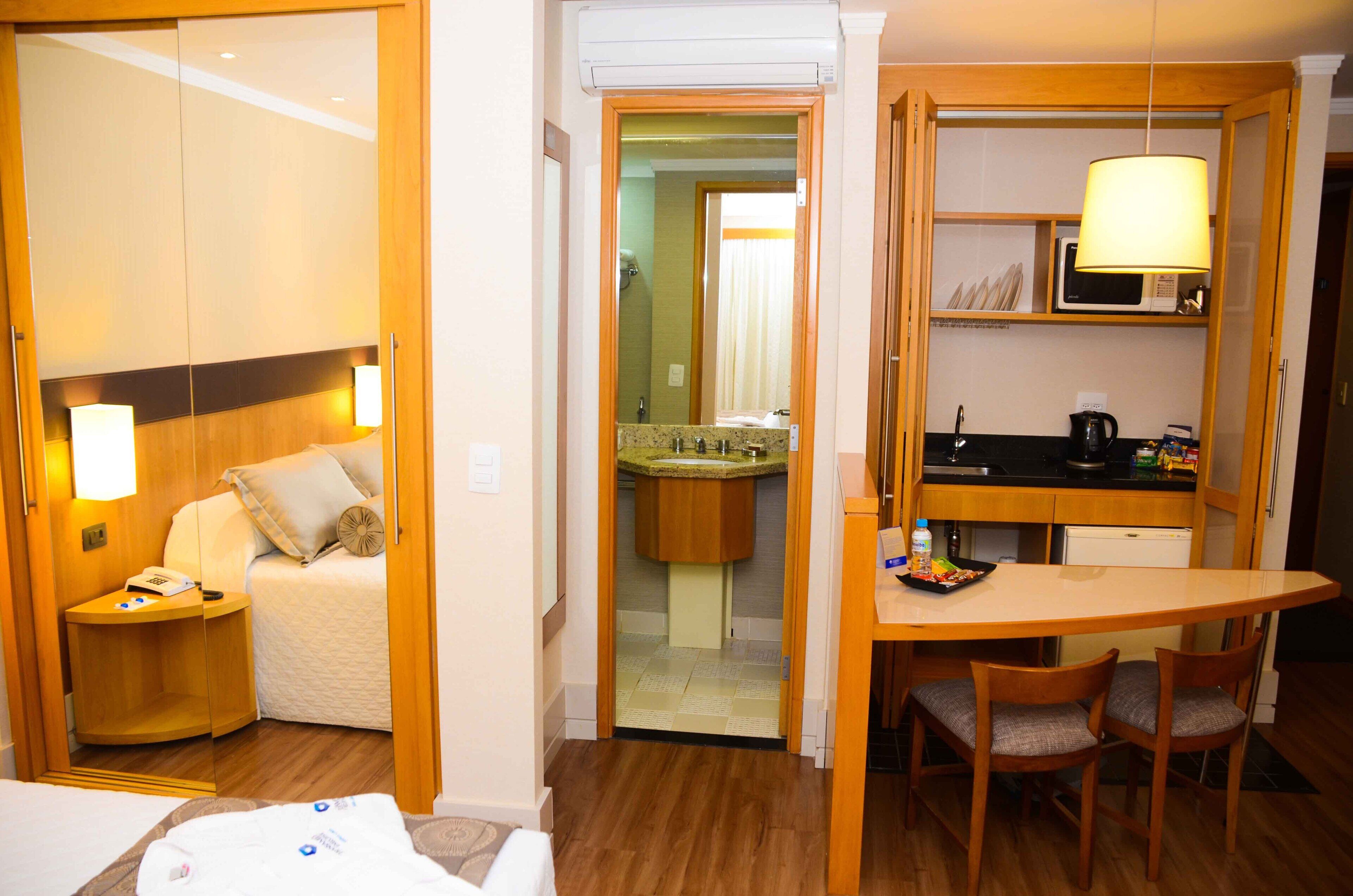 A hotel room with a bed, bedside table, lamp, and a small kitchenette with a microwave, sink, and a table with chairs.