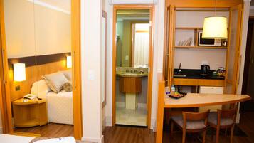 Habitación de hotel con cama, mesita de noche, lámpara y una pequeña cocina americana con microondas, lavamanos y una mesa con sillas.