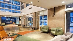 Un hall d’entrée spacieux, avec un grand canapé gris, une petite table ronde et un fauteuil vert. On y trouve plusieurs options d’assises, ainsi qu’une grande fenêtre offrant une vue sur l’extérieur.