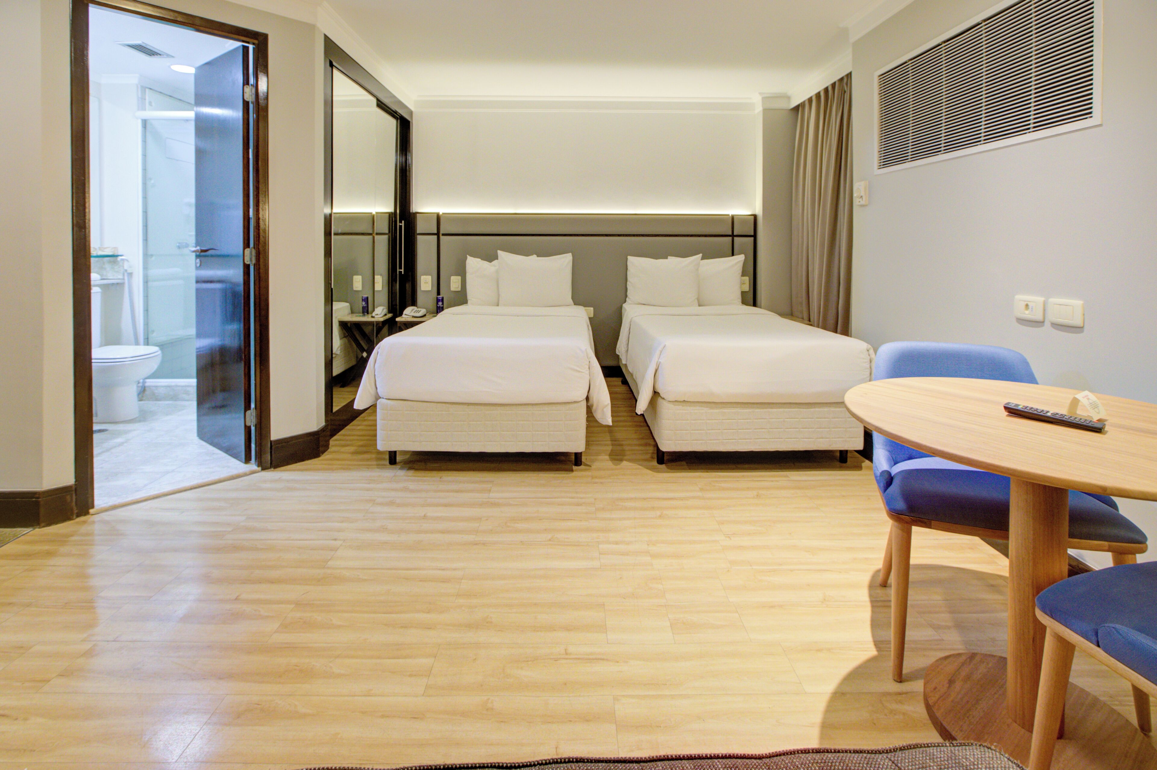 A hotel room with two beds, a bathroom, a small table with a chair, and a window with blinds.